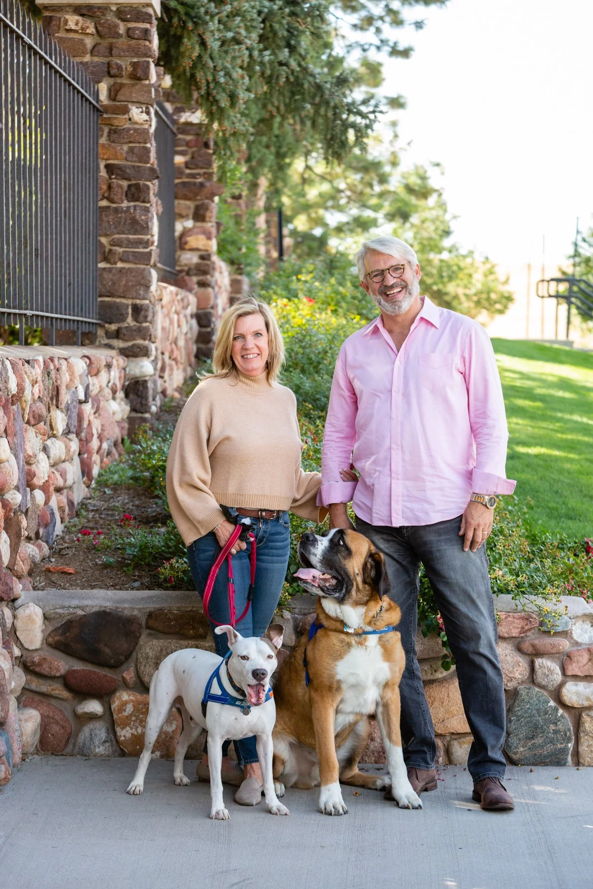 A smiling older couple standing outdoors with three dogs, a man in a pink shirt, a woman in a beige sweater, and two dogs, one large and brown, one white with black spots, on a concrete sidewalk near a stone wall with greenery and trees in the background.