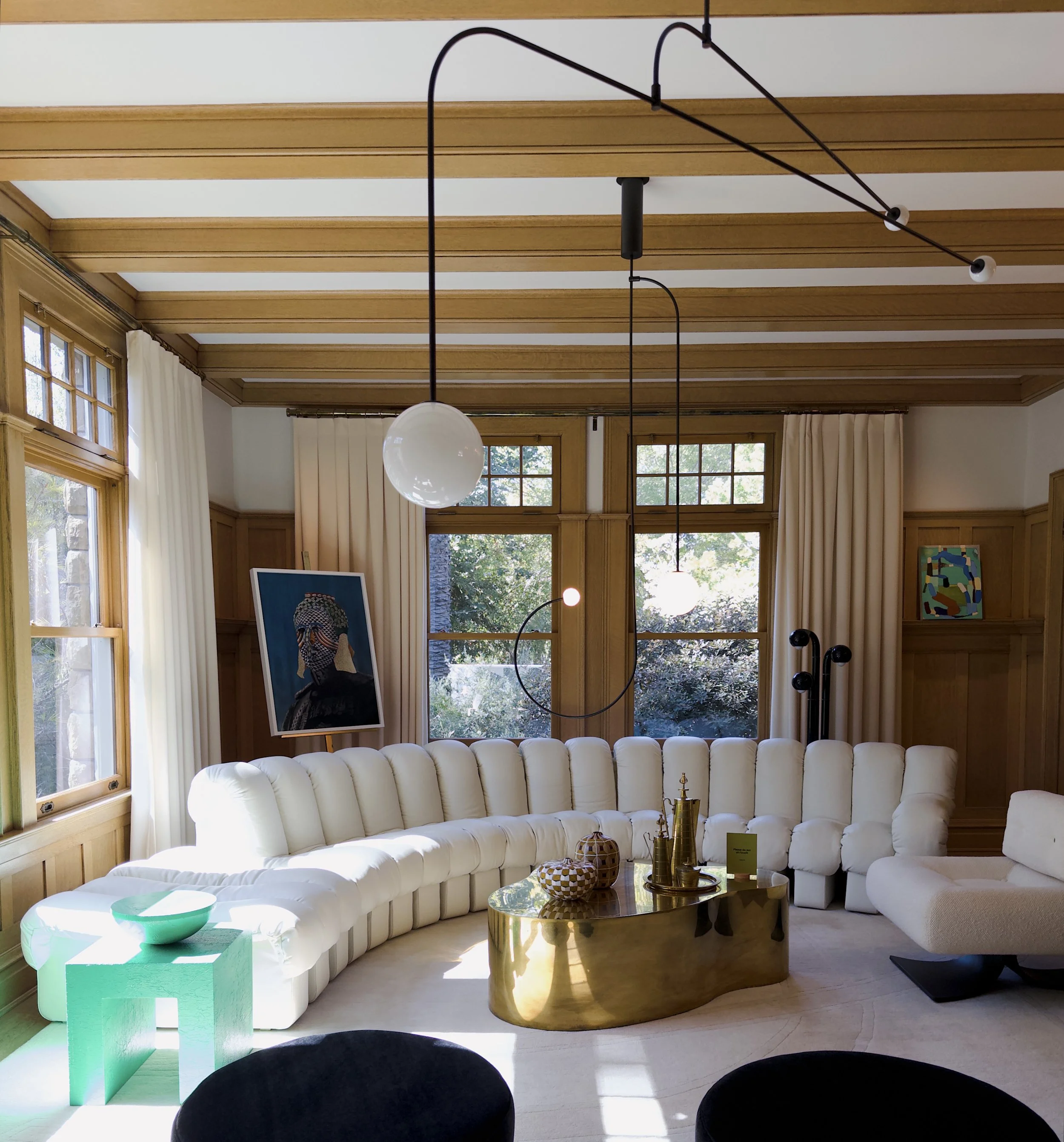 Living room with large cream-colored sofa, wooden coffee table with gold finish, decorative vases, and artworks, surrounded by tall windows with cream curtains and a ceiling with wooden beams and modern black lighting fixtures.