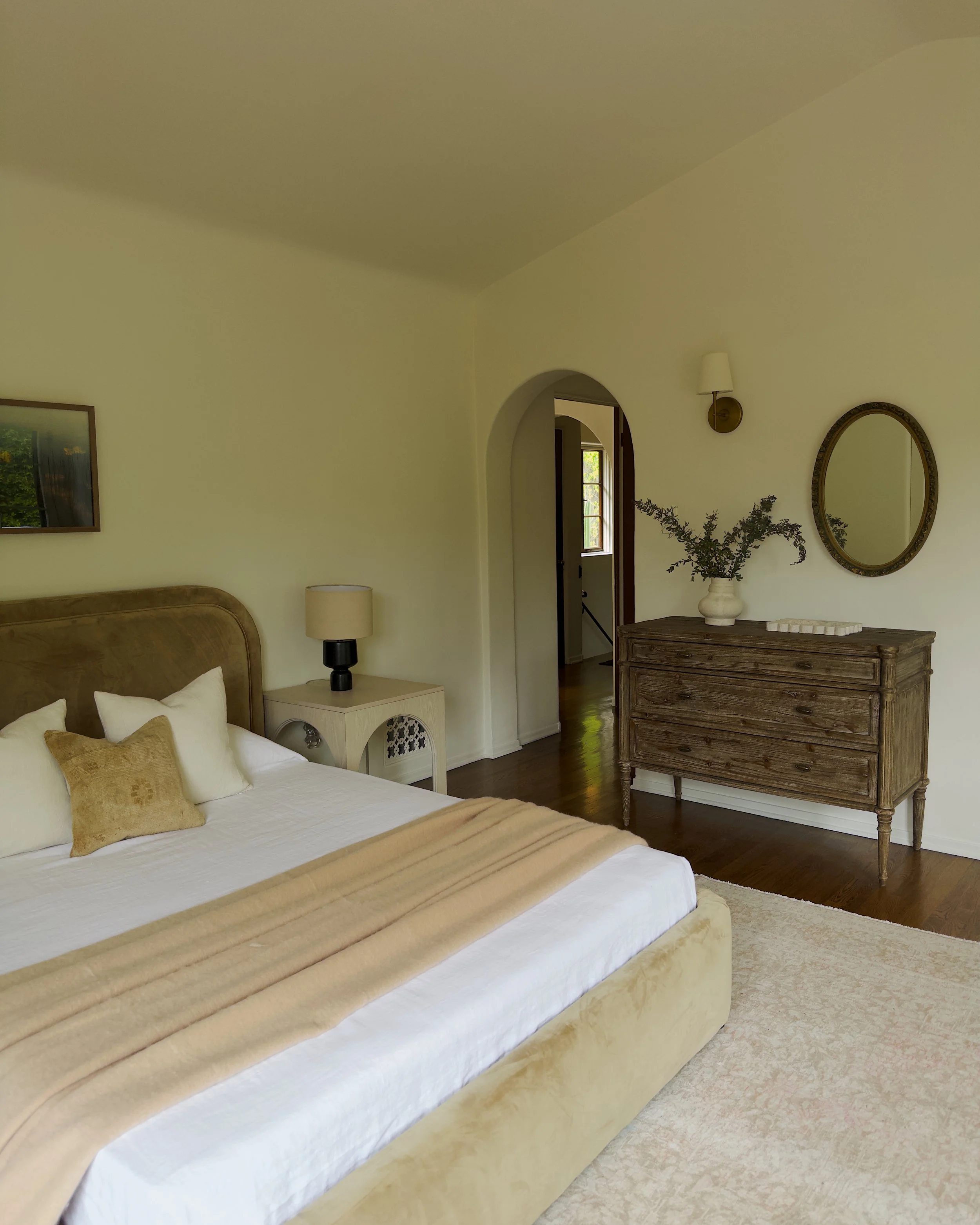 A bedroom with a bed with white bedding and beige pillows, a wooden dresser with a vase of greenery, a mirror, and a wall lamp. An arched doorway leads to a room with a window.
