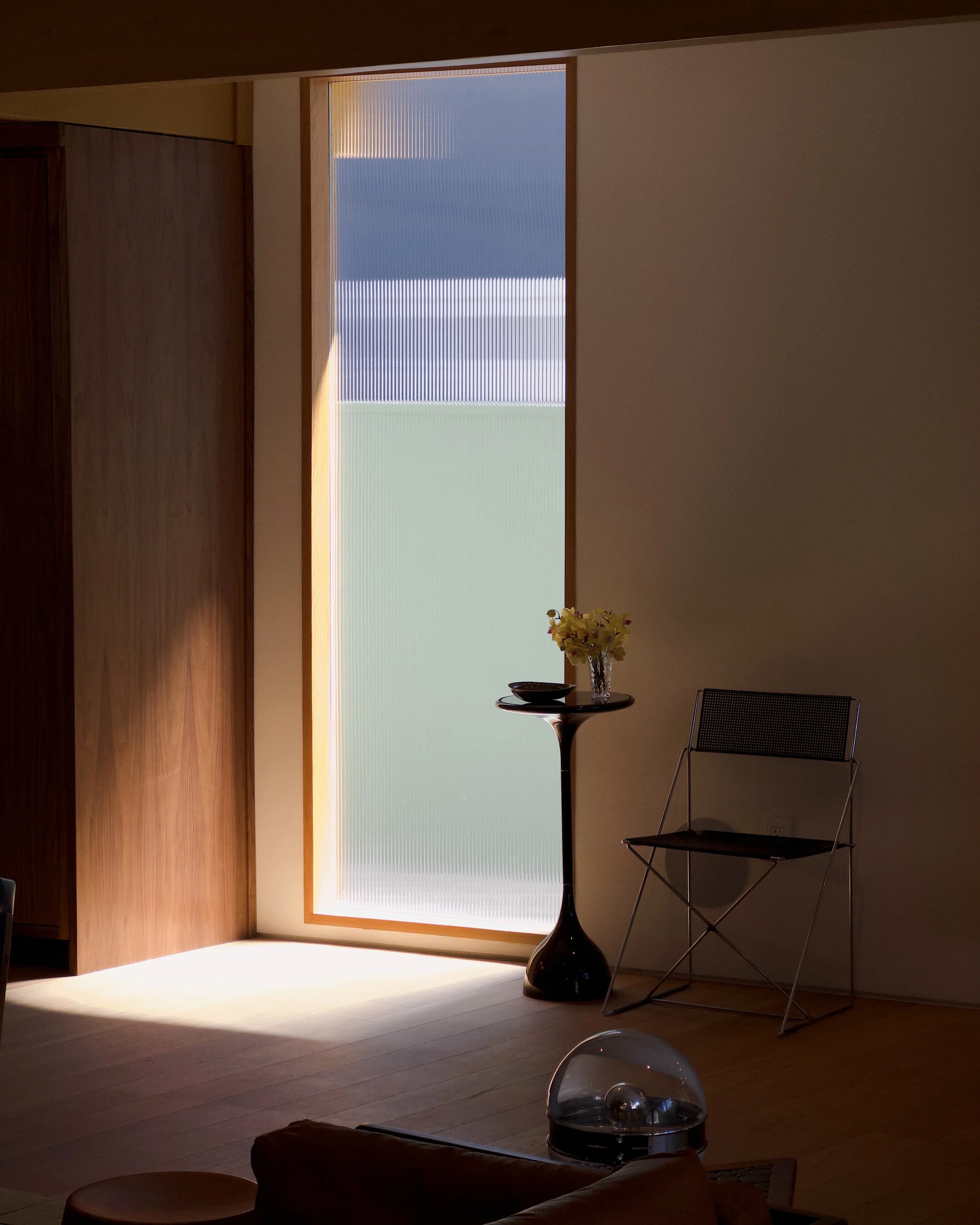 Sunlight coming through textured window blinds illuminating a corner with a small round table holding a decorative vase with flowers and a bowl, next to a modern chair and a floor lamp, in a room with wooden floors and minimal decor.