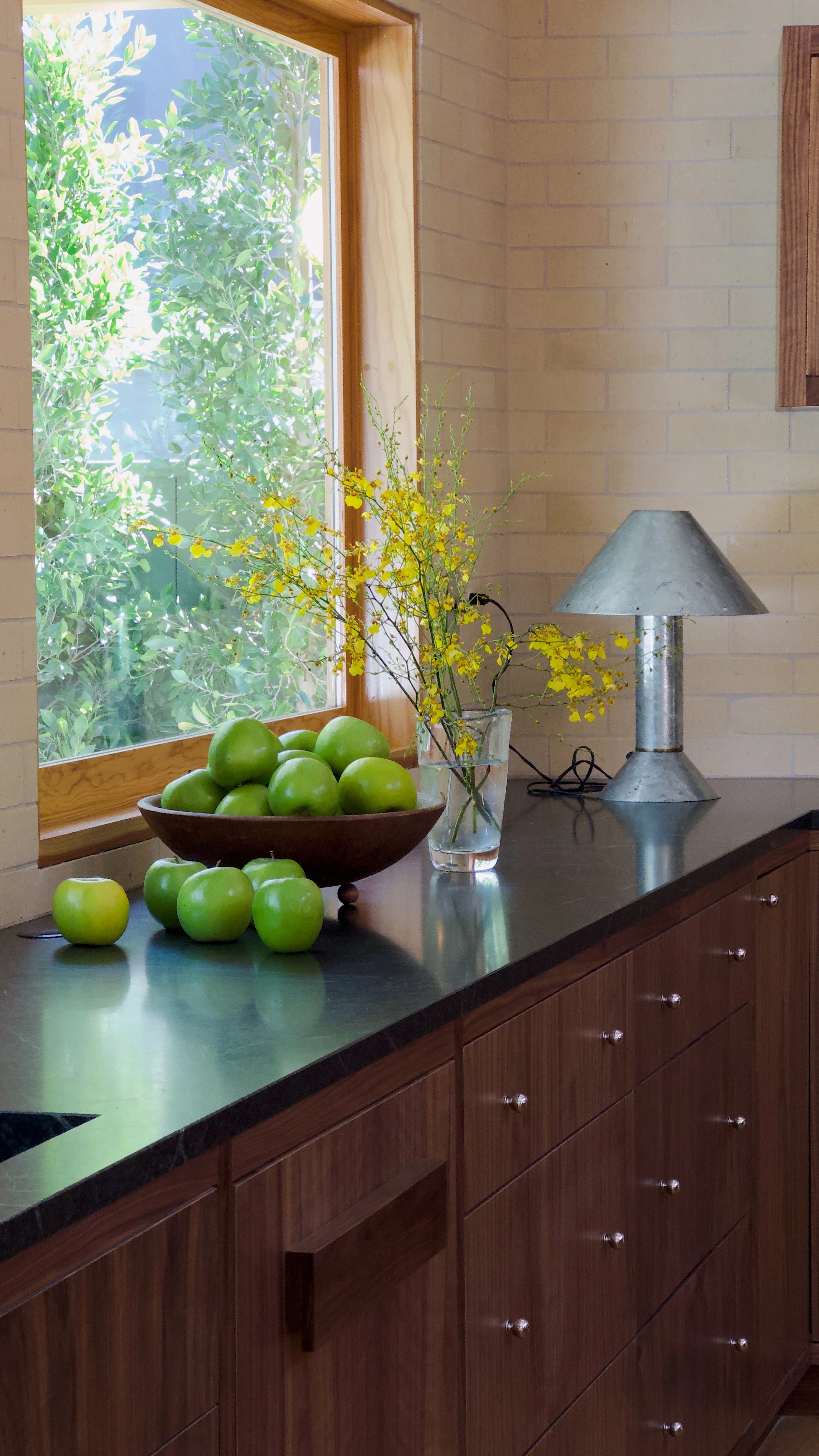 Green apples in a wooden bowl, a few scattered on a dark countertop, a window with greenery outside, a vase with yellow flowers, and a metallic table lamp on a kitchen counter.