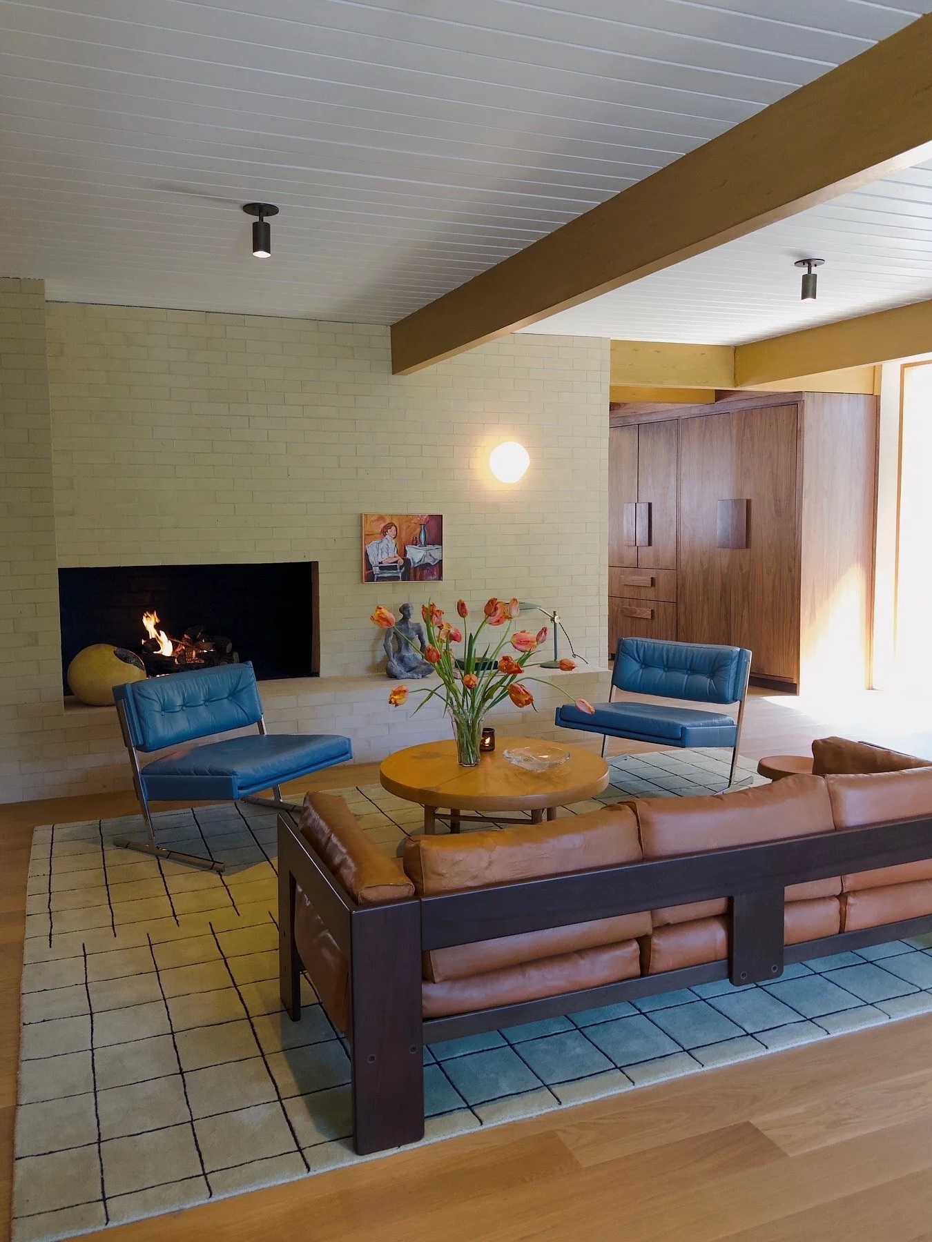 Living room with a beige brick fireplace, two blue armchairs, a round wooden coffee table with a vase of orange tulips, a modern brown leather sofa, a patterned area rug, and wooden beams on the ceiling. There's also a lamp, a small sculpture, and a 