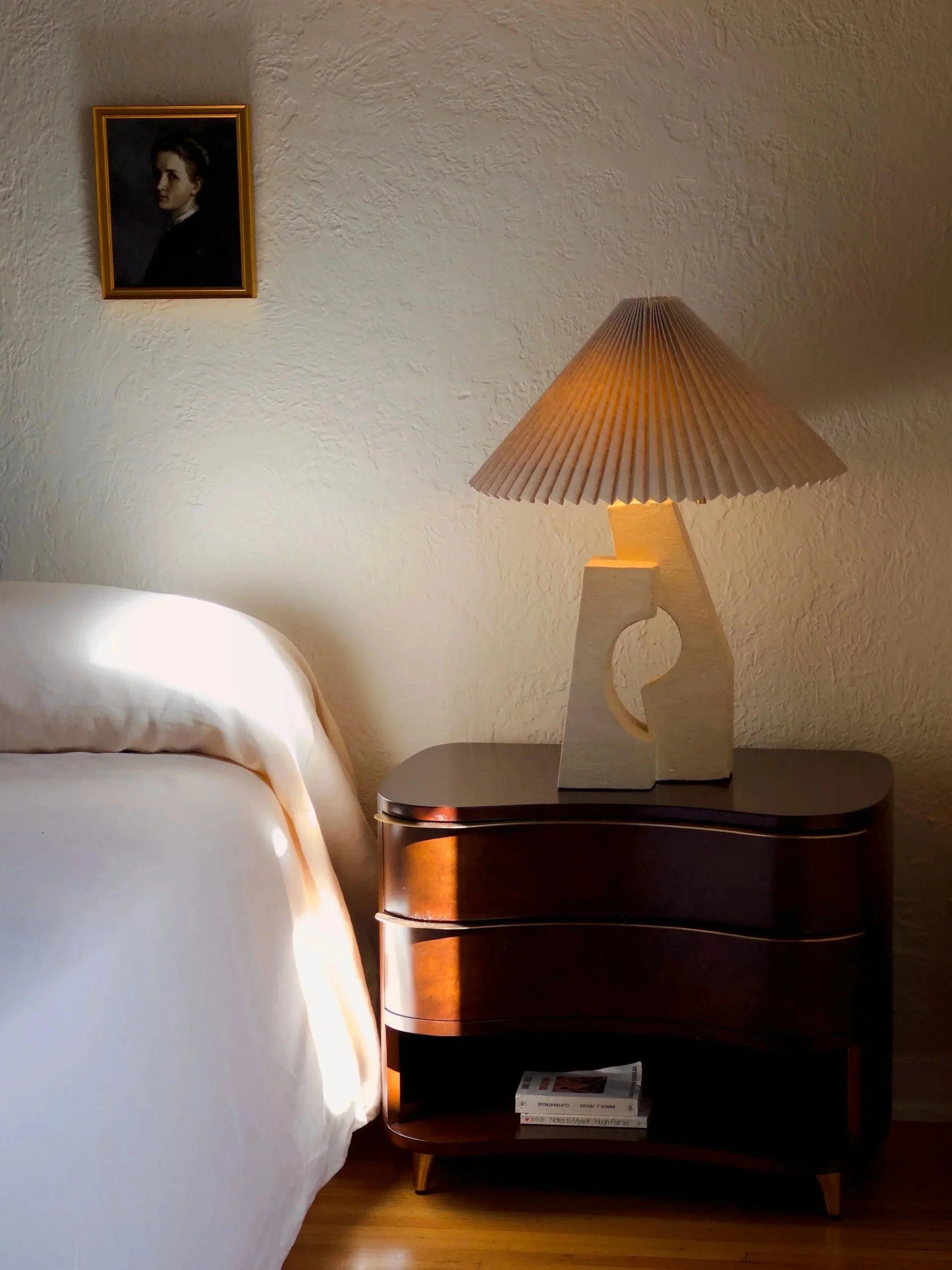 A bedroom with a white bed, a wooden nightstand with a closed shelf, a table lamp with a pleated shade, and a framed portrait of a woman on the textured wall.
