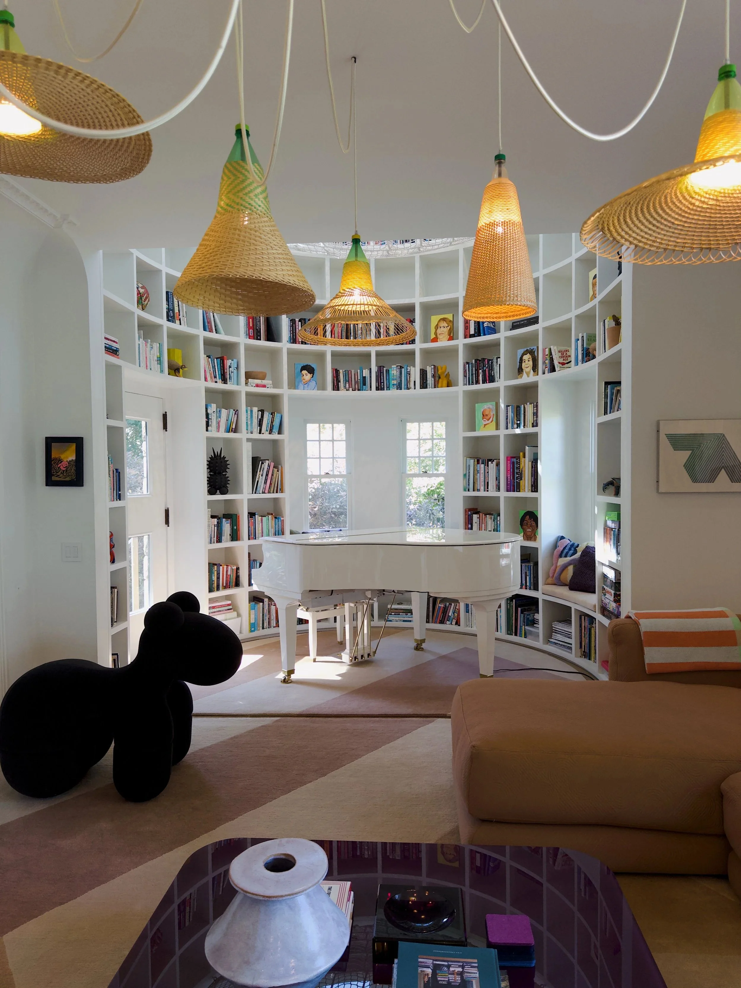 Living room with a white grand piano, a curved white bookshelf filled with books and art, hanging wicker pendant lights, a beige armchair with striped pillow, a black sculpture, and a glass coffee table with a white lamp, books, and small decorative 