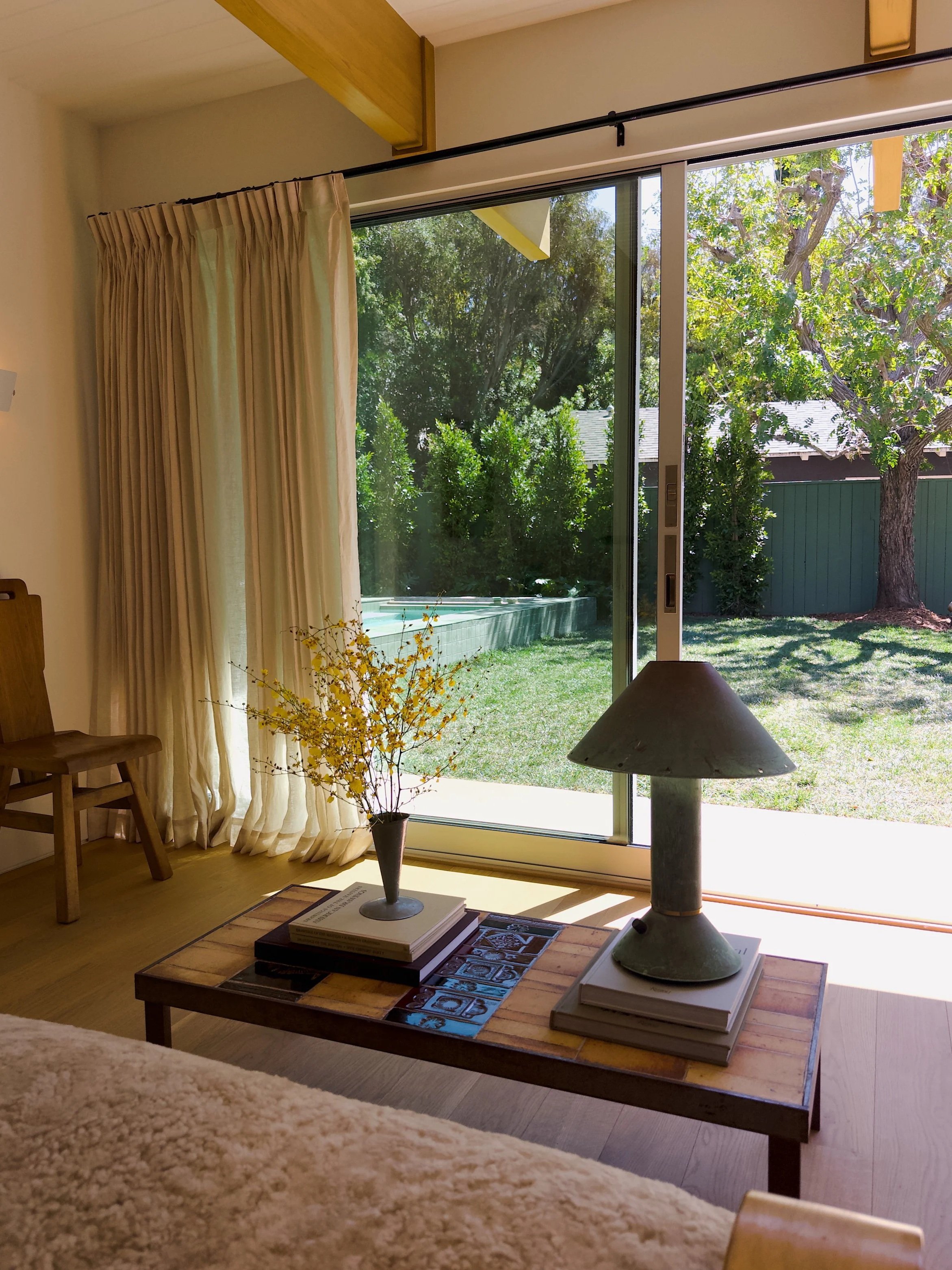 Living room with large glass sliding door showing green trees outside. Inside there is a wooden coffee table with books and a lamp, a small bouquet of yellow flowers, and a wooden chair. Cream-colored curtains partially cover the door.
