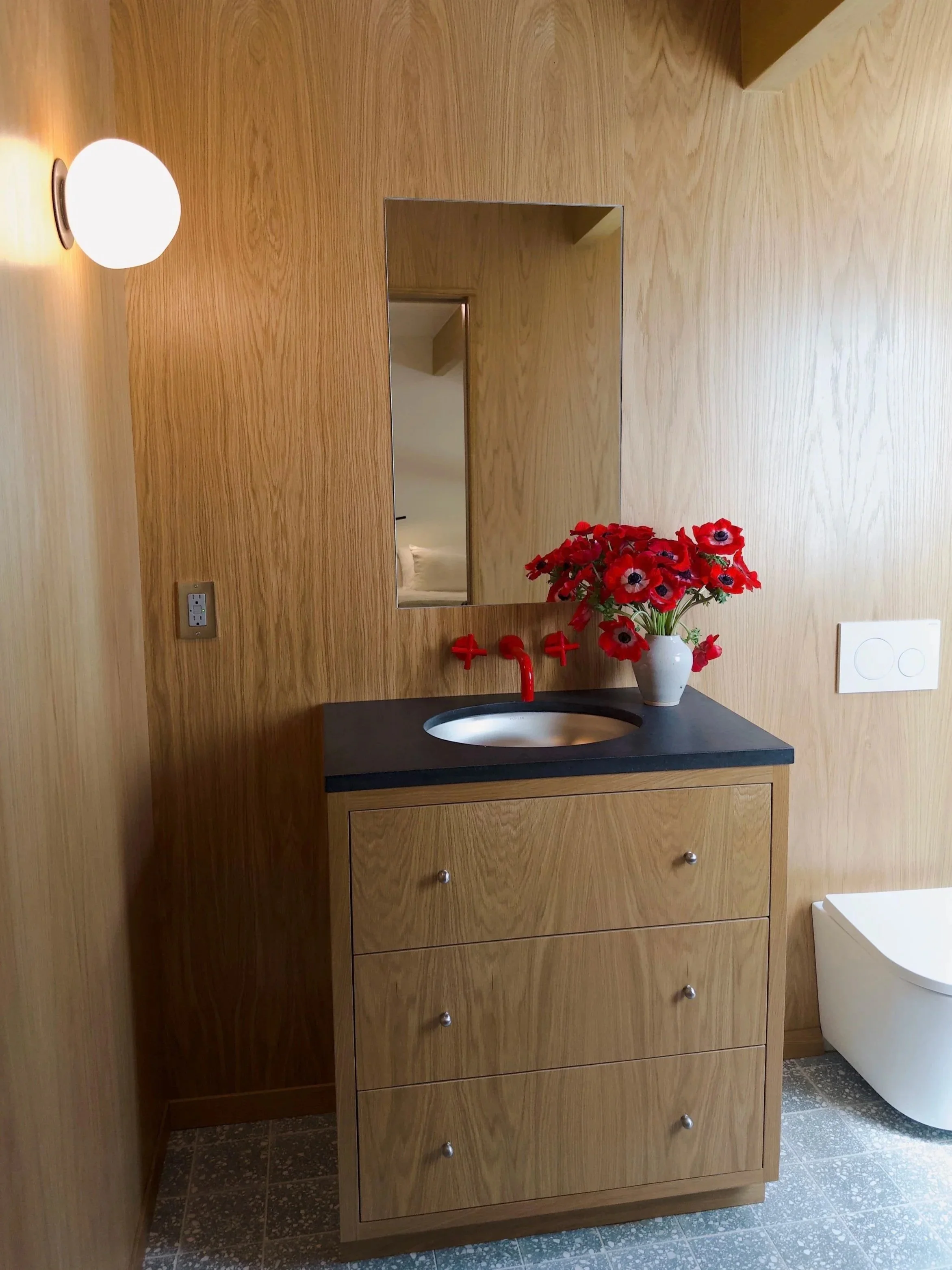 A wooden bathroom cabinet with a black countertop and a round sink, topped with a vase of red flowers. A mirror is mounted above the cabinet, reflecting part of a bed in an adjacent room. A wall-mounted light fixture is on the left, and a white toile
