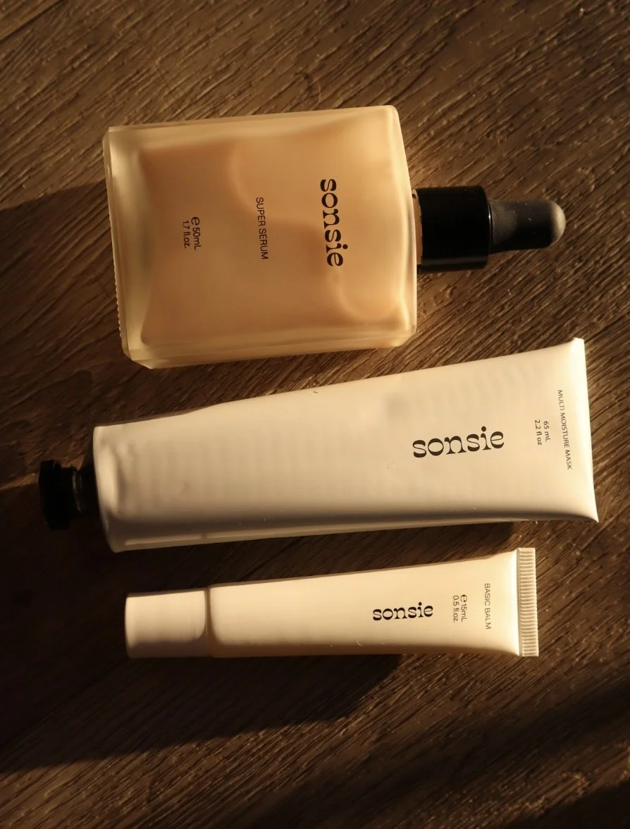 Three skincare products from the brand Sonsie placed on a wooden surface. The top bottle is a 50ml super serum with a dropper cap, the middle tube is a 65ml multi-moisture mask, and the bottom tube is 5ml of basic balm.