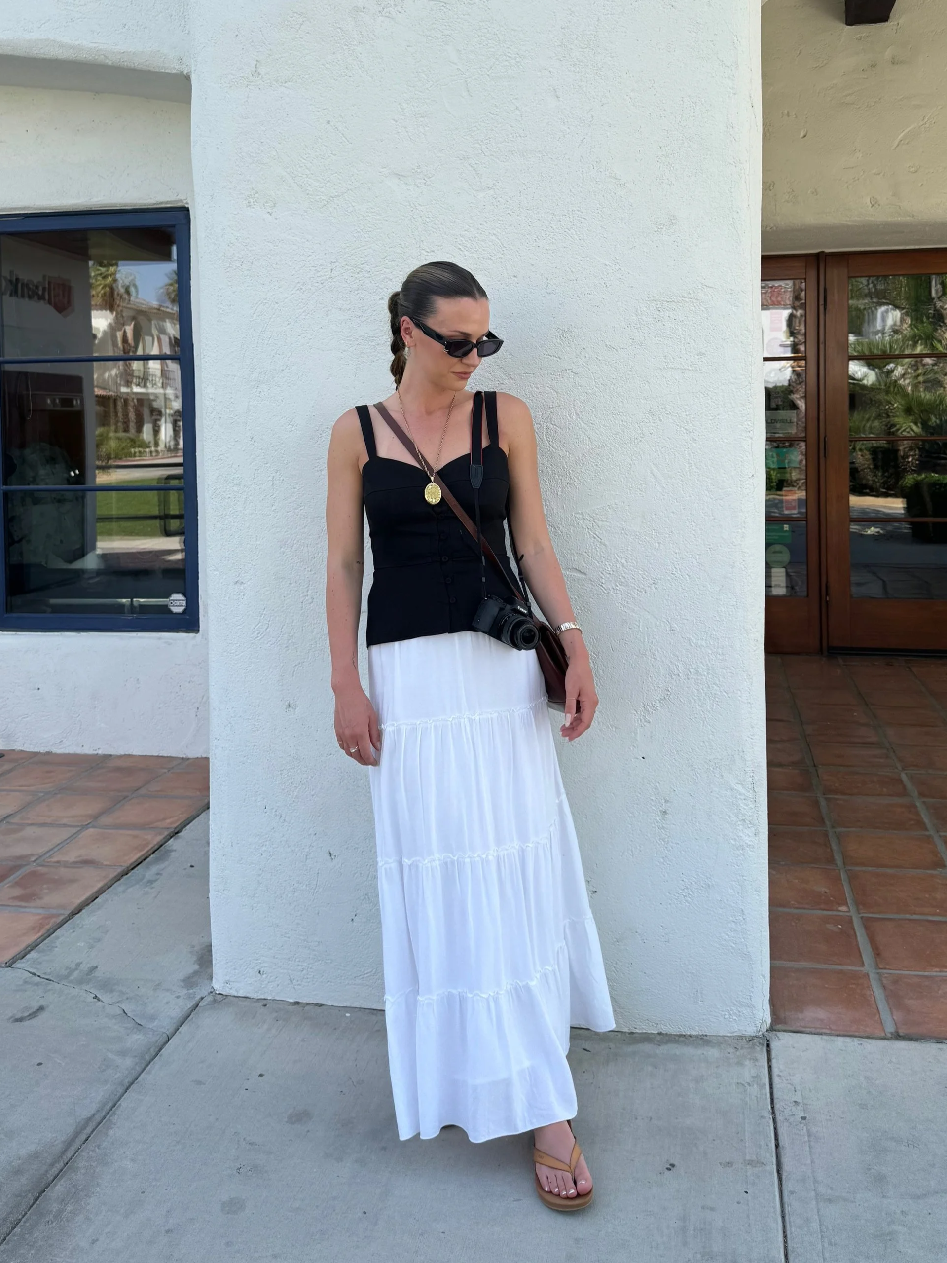 A woman in black sunglasses, a black tank top, a white pleated skirt, and tan sandals stands outside in front of a white wall, with a camera hanging from her neck and a necklace with a pendant.