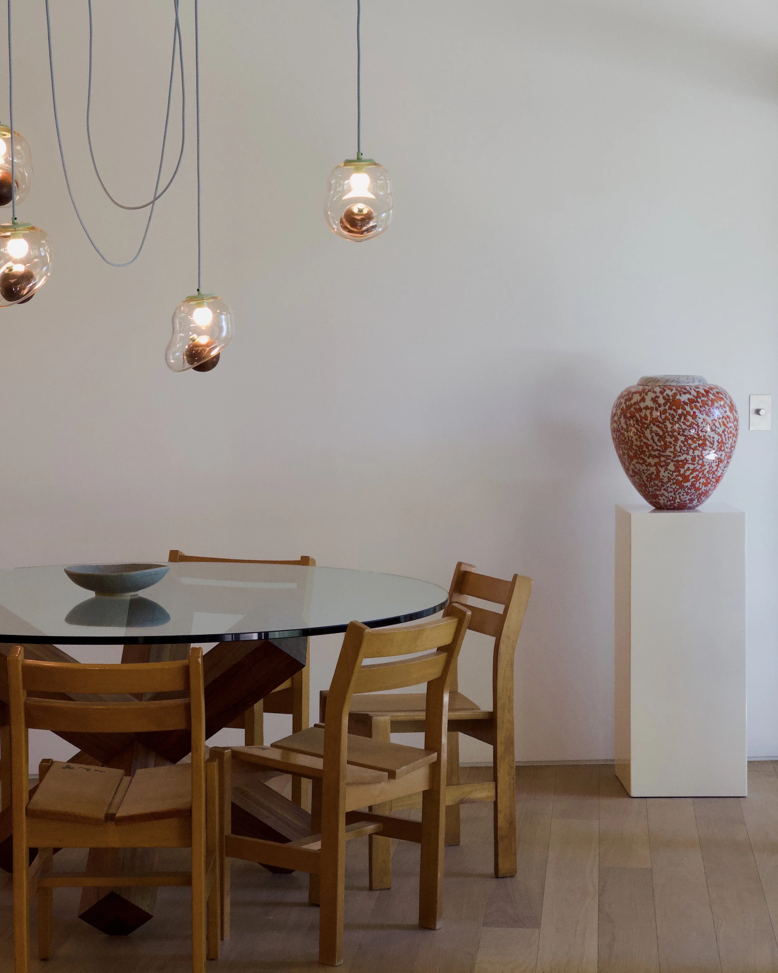 A dining table with wooden chairs, a decorative ceramic vase, and hanging glass pendant lights in a minimalist room.