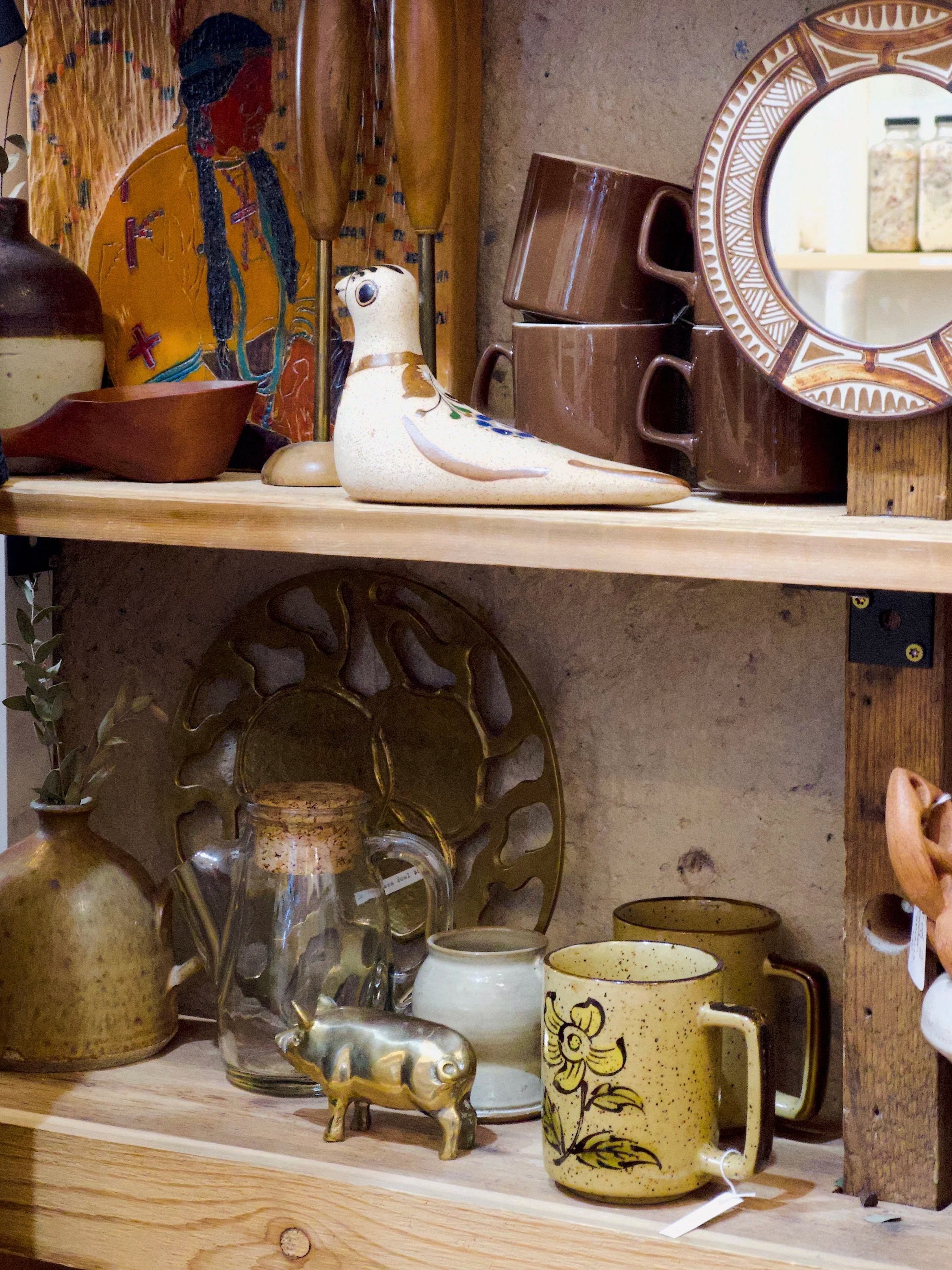 A collection of various decorative items on a wooden shelf, including ceramic and glass mugs, a gold-colored animal figurine, a small white ceramic vessel, a greenish glass vase, and a ceramic plate with intricate patterns. In the background, there is a framed picture of a woman and a mirror.