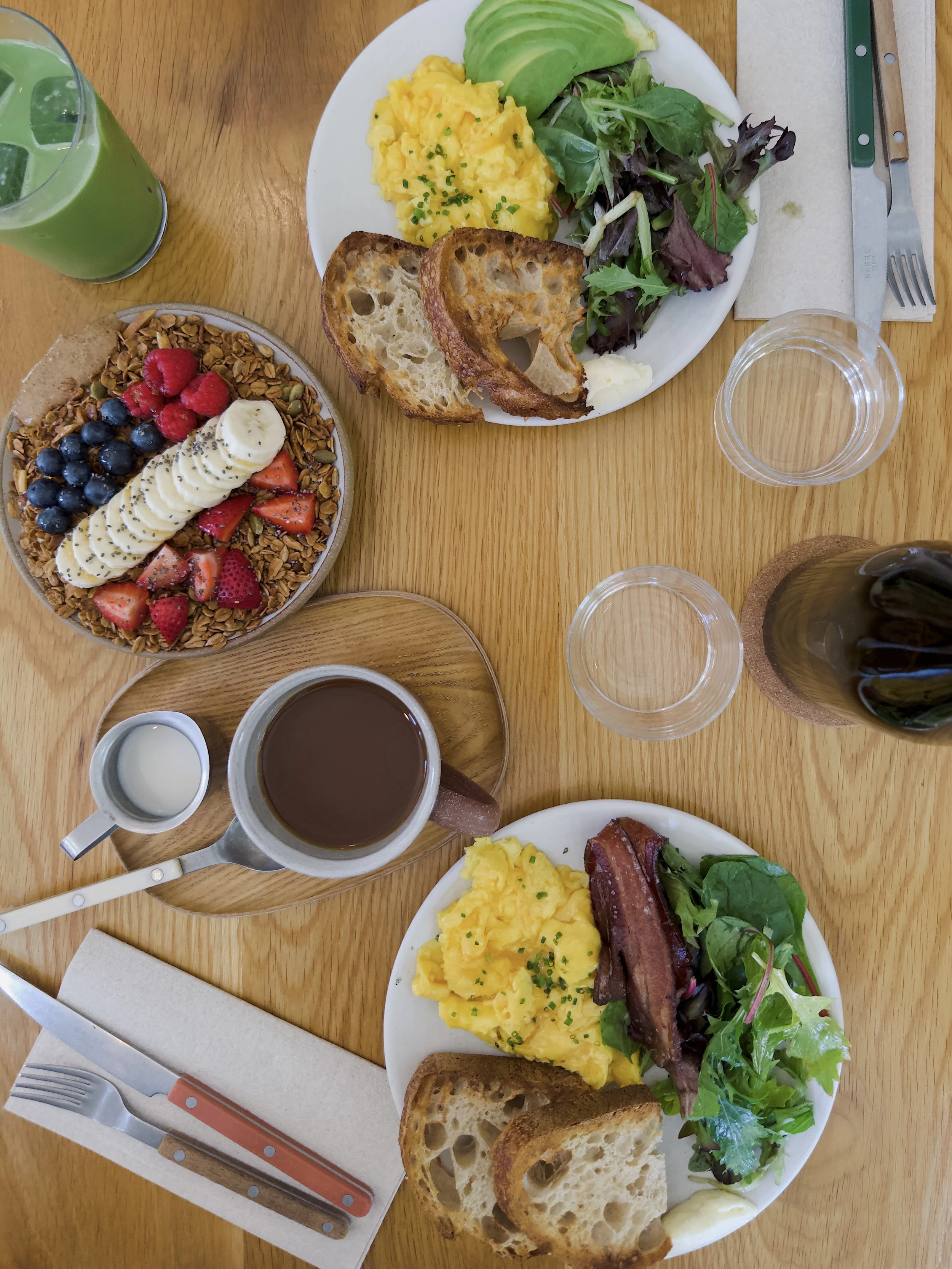 A breakfast meal with scrambled eggs, fresh greens, toasted bread, and bacon on a white plate. A side of fruit with strawberries, blueberries, and bananas, garnished with seeds. A green beverage with ice in a glass, and a cup of coffee. Silverware on