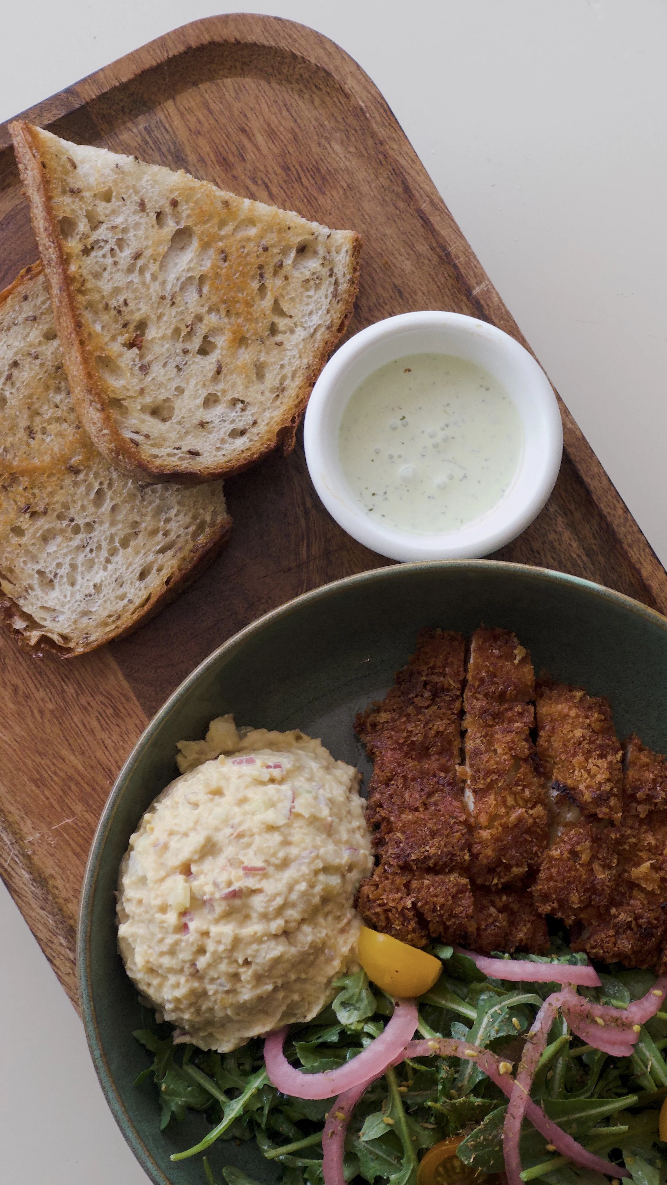 Slices of toasted bread, a small cup of dressing or dip, part of a salad with fried chicken, potato salad, and fresh greens with sliced onions and cherry tomatoes.