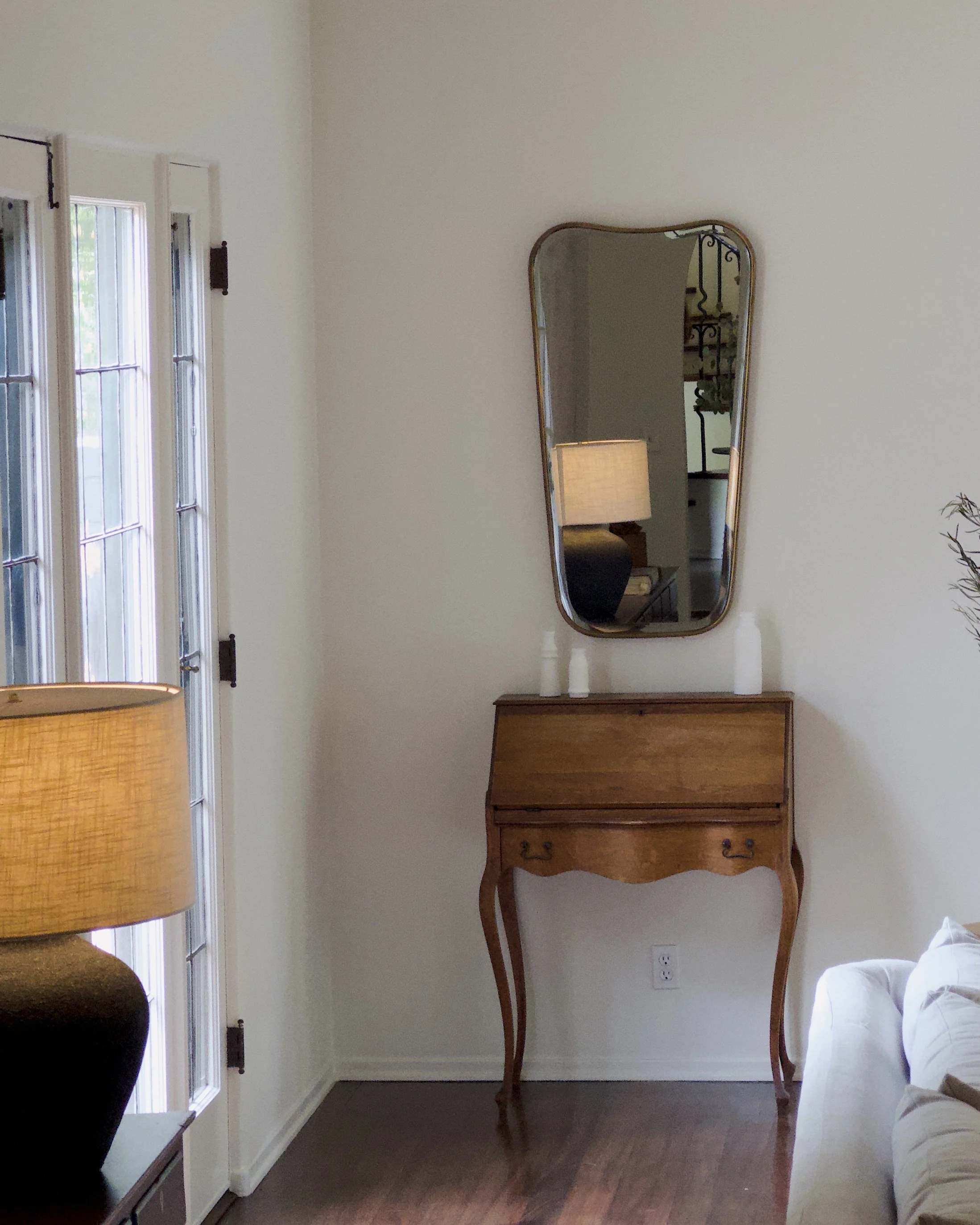 A corner of a room featuring a vintage wooden writing desk with curved legs, a larger oval mirror above it, a small table with candles beneath the mirror, a window with vertical blinds on the left, a beige wall, a lamp with a fabric shade on a side t