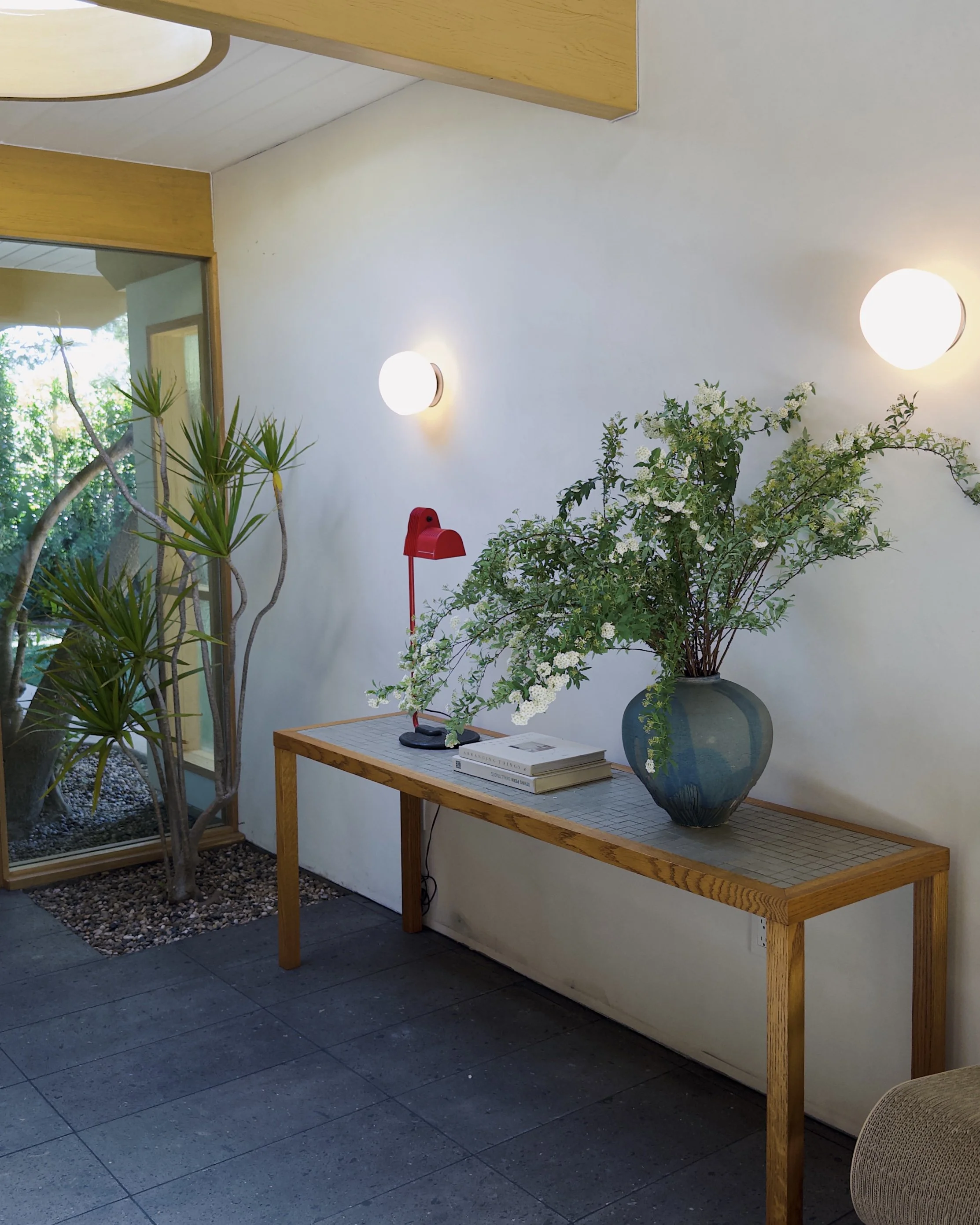 Interior view of a modern room with a wooden console table decorated with a large ceramic vase filled with white flowering greenery, a red desk lamp, and books. The room has white walls with two spherical wall-mounted lights, large windows with a vie