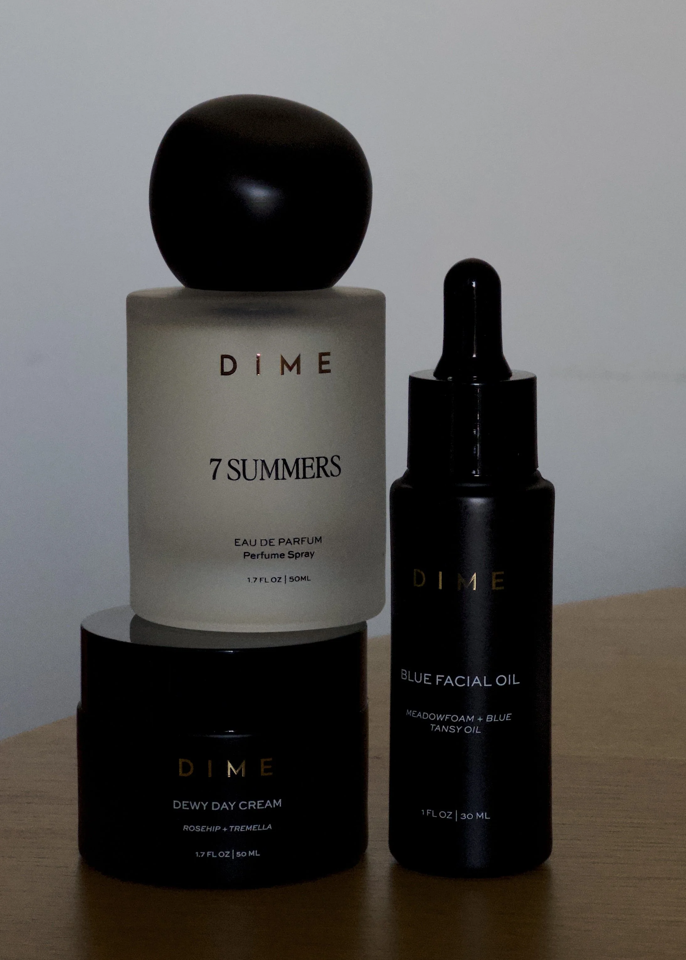 Three skincare or cosmetic products on a wooden surface: a white bottle labeled 'DIME 7 SUMMERS,' a black bottle with a dropper labeled 'DIME BLUE FACIAL OIL,' and a black container labeled 'DIME DEWY DAY CREAM.'