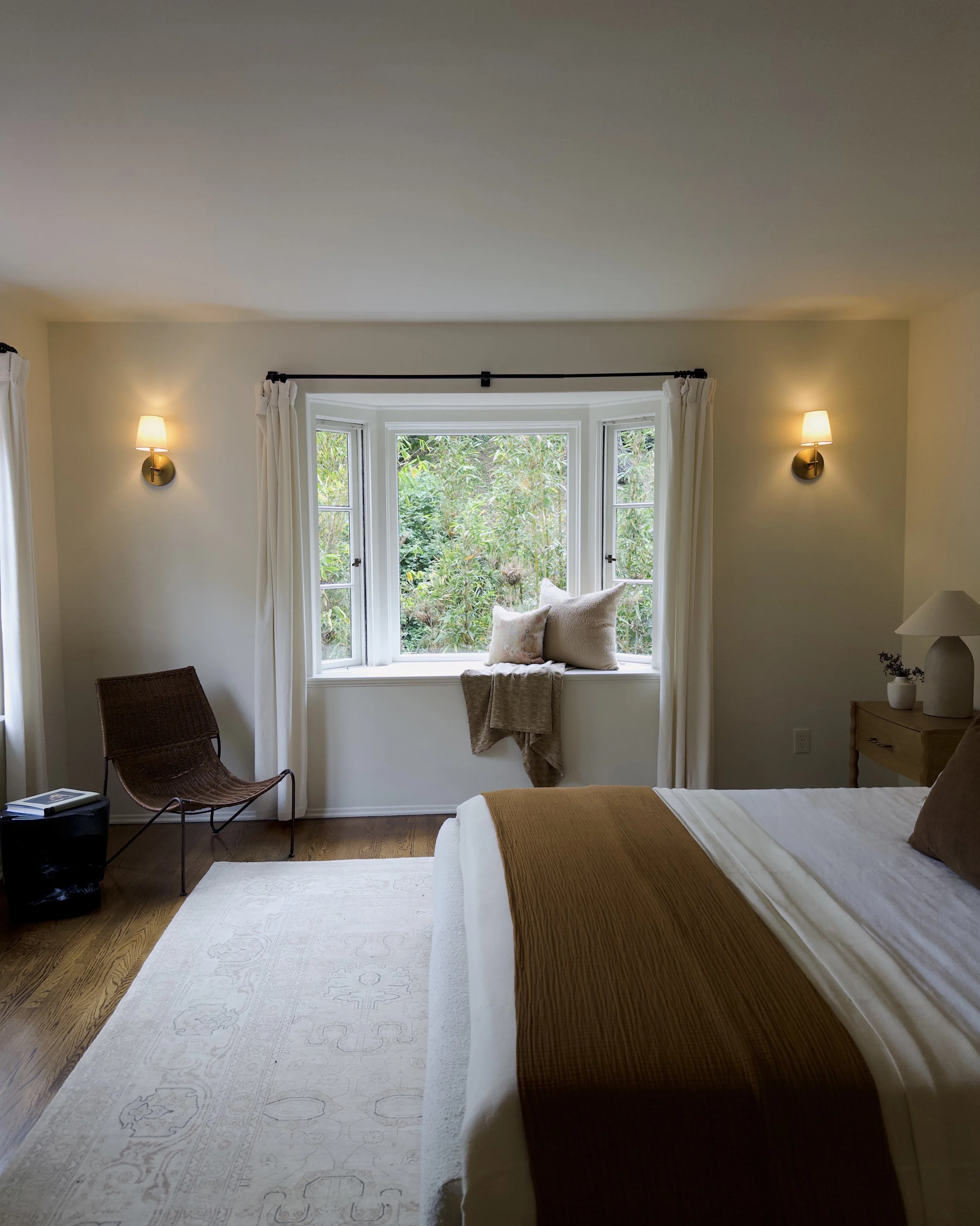 A bedroom with a white bed, mustard yellow blanket, brown pillows, and a large white area rug. There is a window with white curtains, a chair, and two wall sconces with lights on.