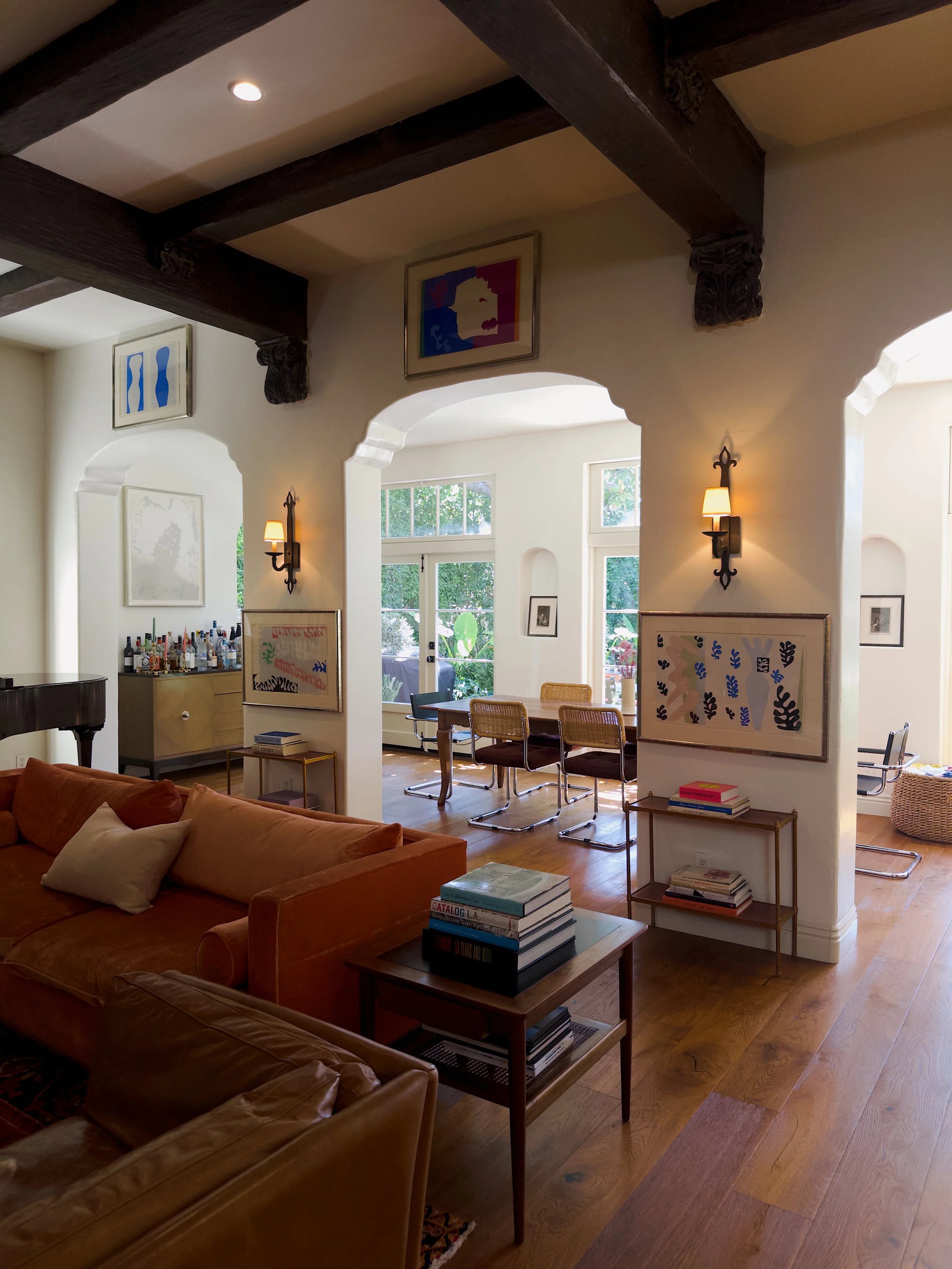 Living room with brown leather sofa, wooden side table with stacked books, artwork on the walls, and a dining table with wicker chairs, illuminated by wall sconces and natural light from windows.