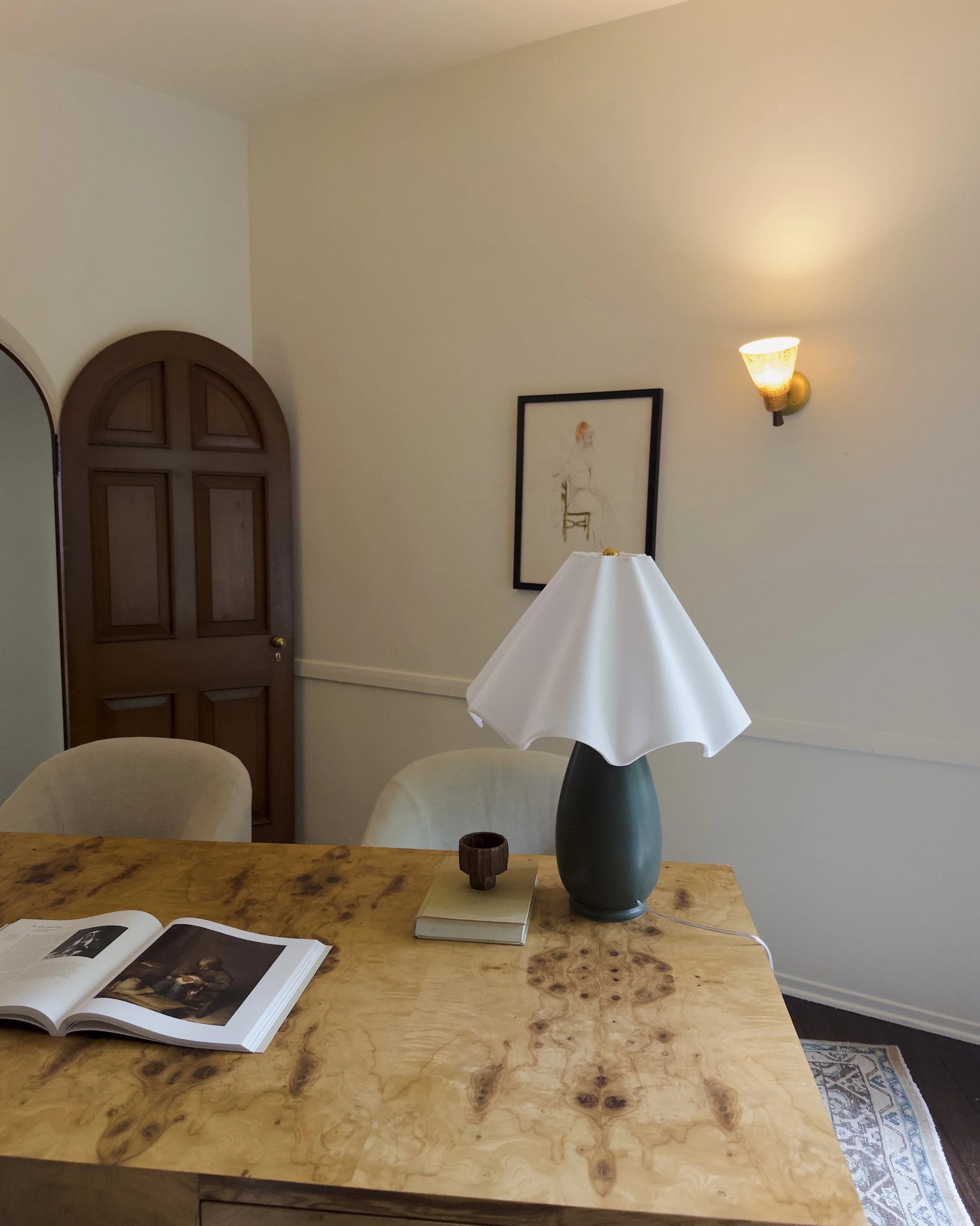 Interior of a room with a wooden table, a lamp with a white shade, a small decorative wooden cup, and a couple of books. There is a wall with a black framed picture, a wall-mounted light fixture, an arched door, and part of a mirror on the left side.