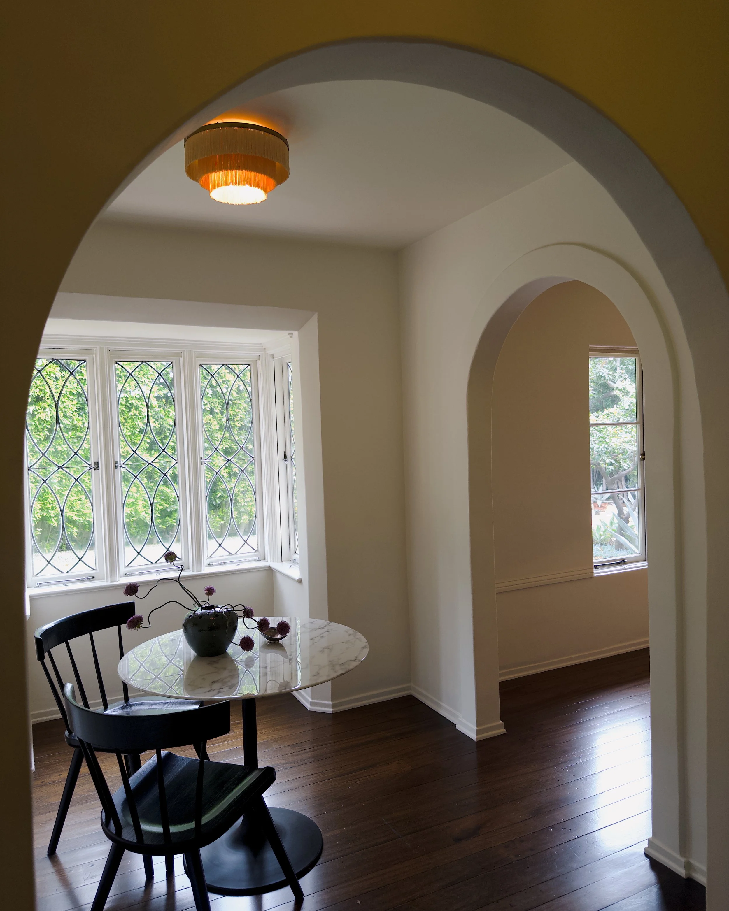 A dining area with a round marble-topped table, two black chairs, a vase with pink flowers, a large window with decorative glass, and a view of green foliage outside.