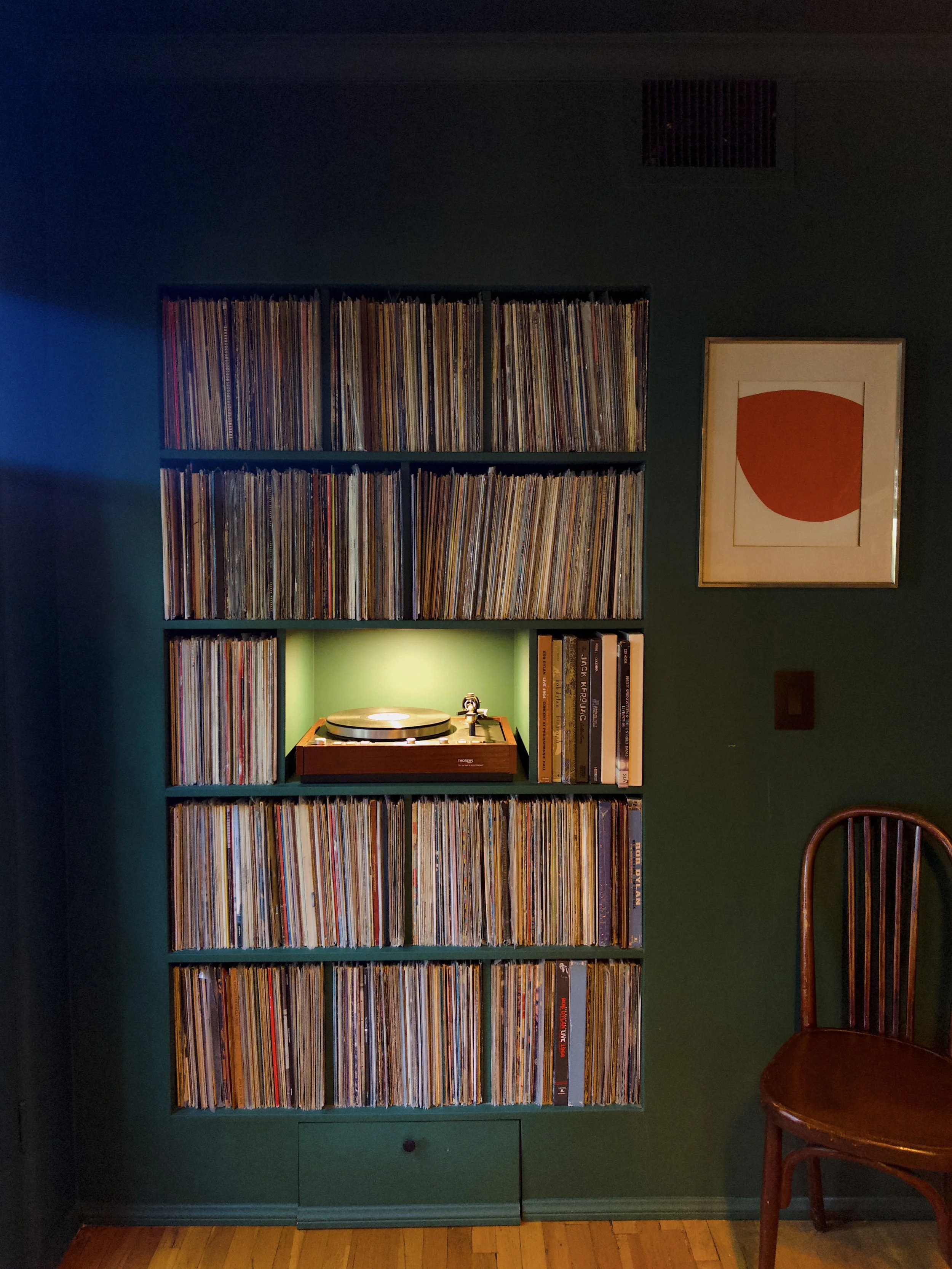 A dark green bookshelf filled with vinyl records, a framed abstract art print, a vintage turntable with a green backlight underneath, and a wooden chair with a curved backrest next to the bookshelf.