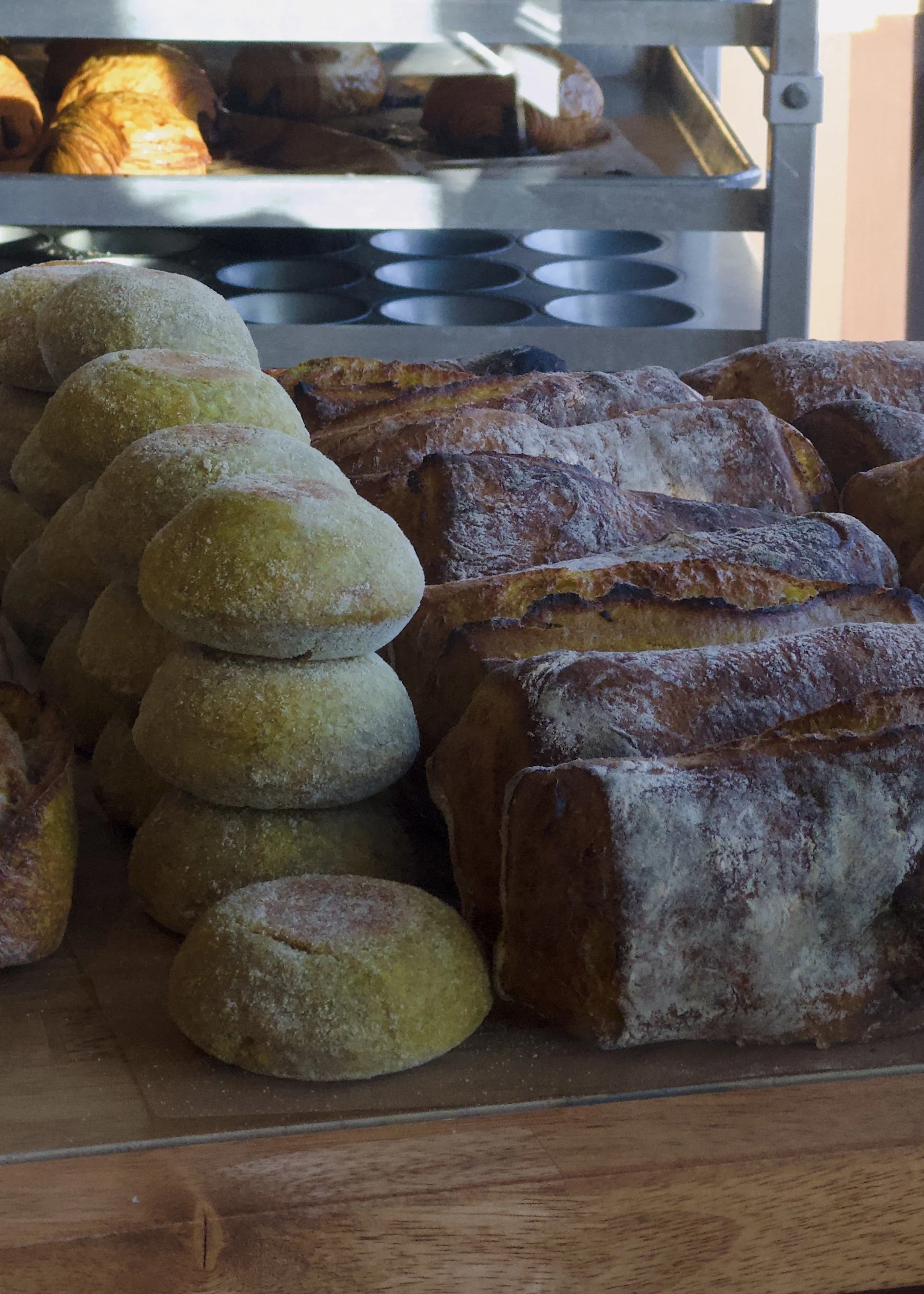 Stacked bread rolls and baguettes on a wooden surface, with a metal bakery display shelf in the background.