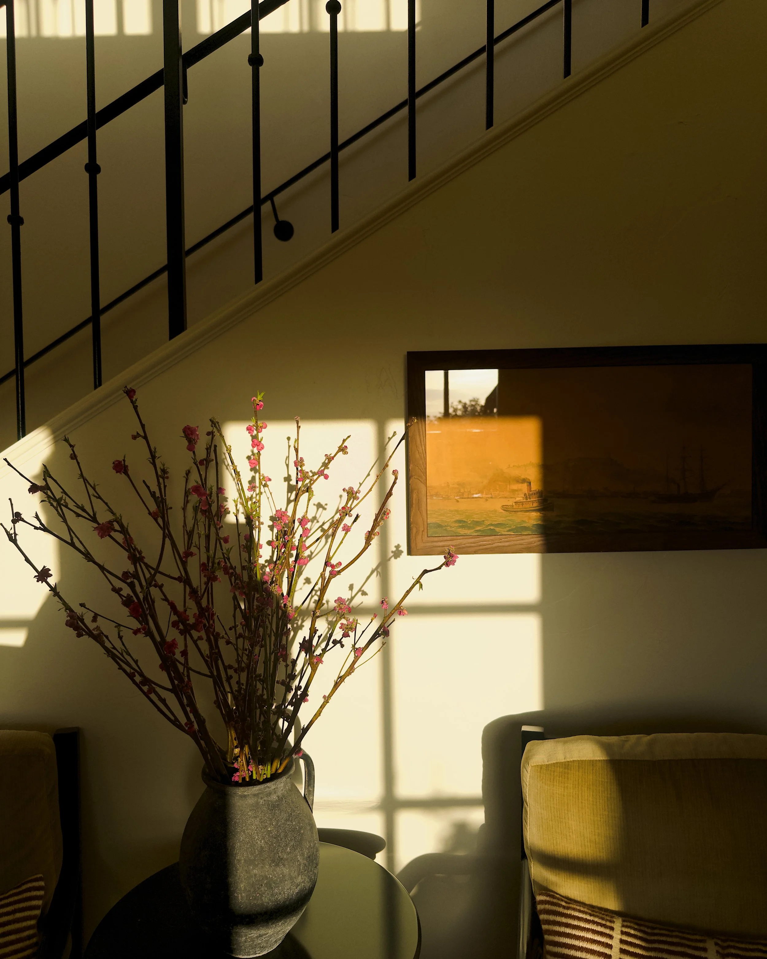 A large grey vase with pink flowering branches inside, placed on a table next to a yellow upholstered chair. Sunlight casts shadows of window panes onto the wall.