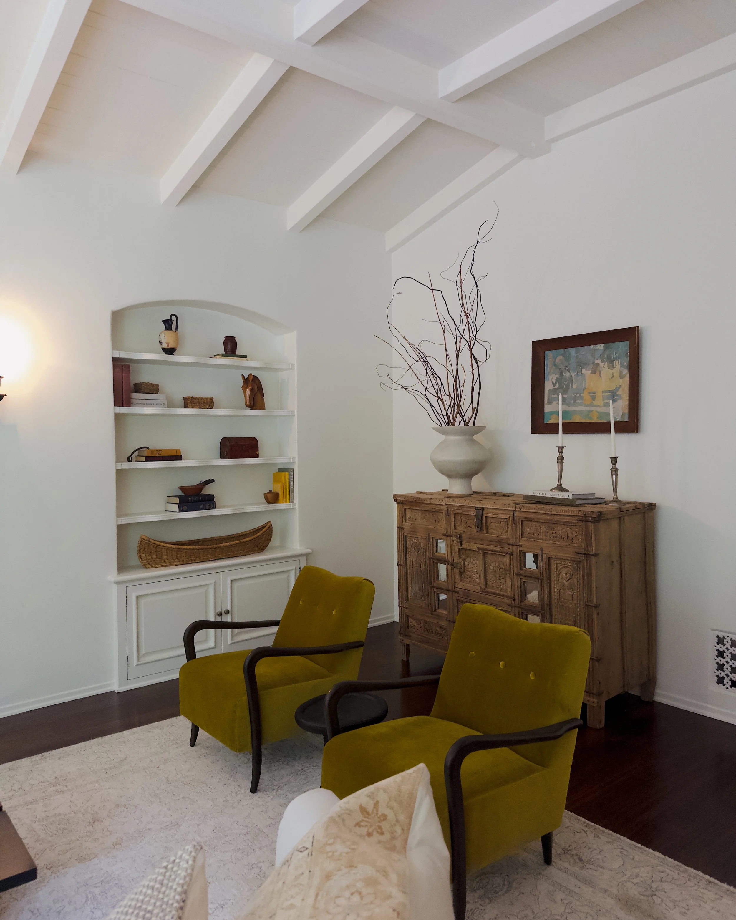Living room with white ceiling beams, a built-in white bookshelf with decorative items, a large wooden sideboard with a floral vase and tall candlesticks, and green armchairs, on a white rug with dark floors.