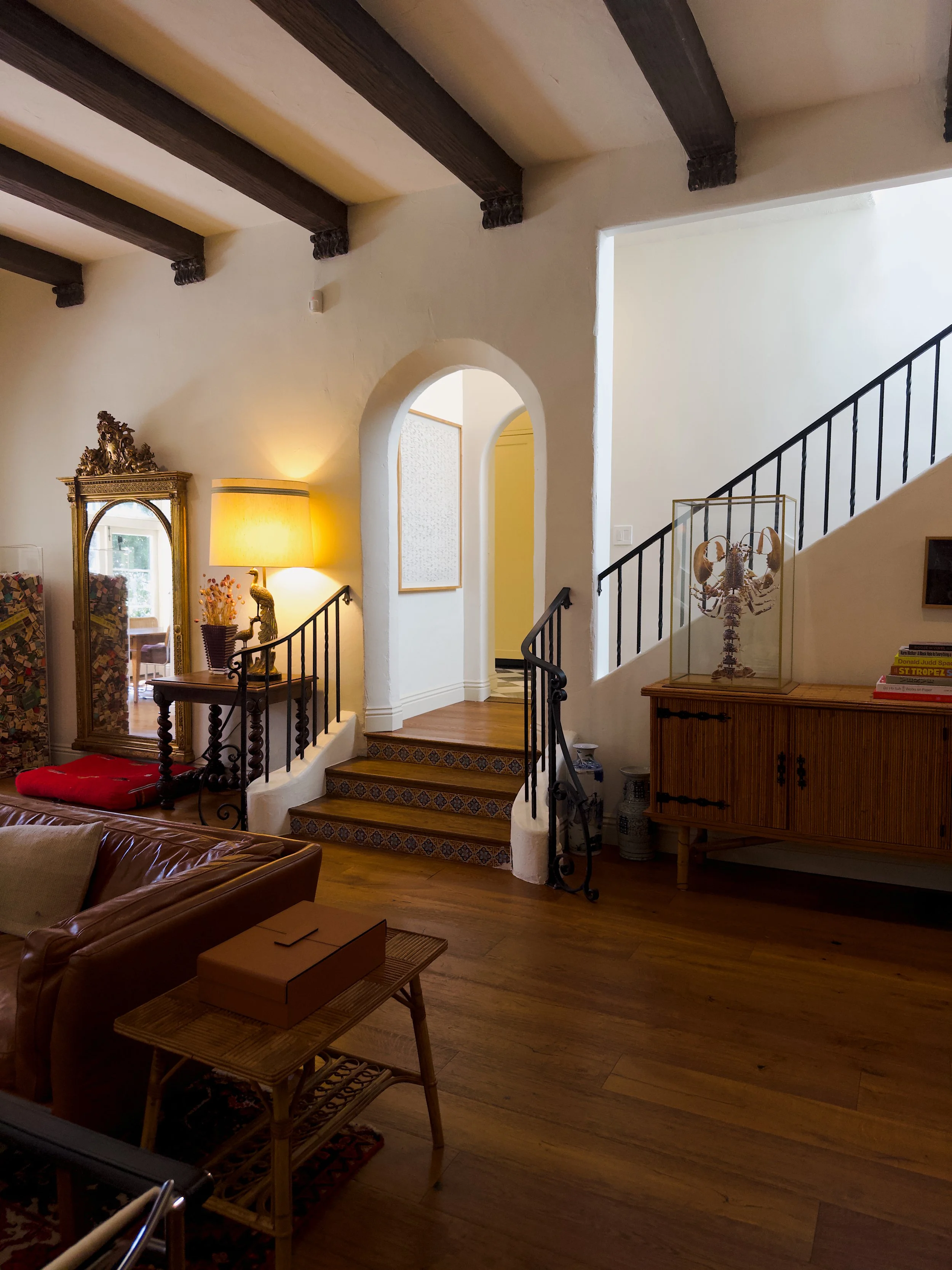 Living room with wooden beams on the ceiling, staircase with black railing, a vintage mirror, a lamp, and a display case with a lobster skeleton inside.