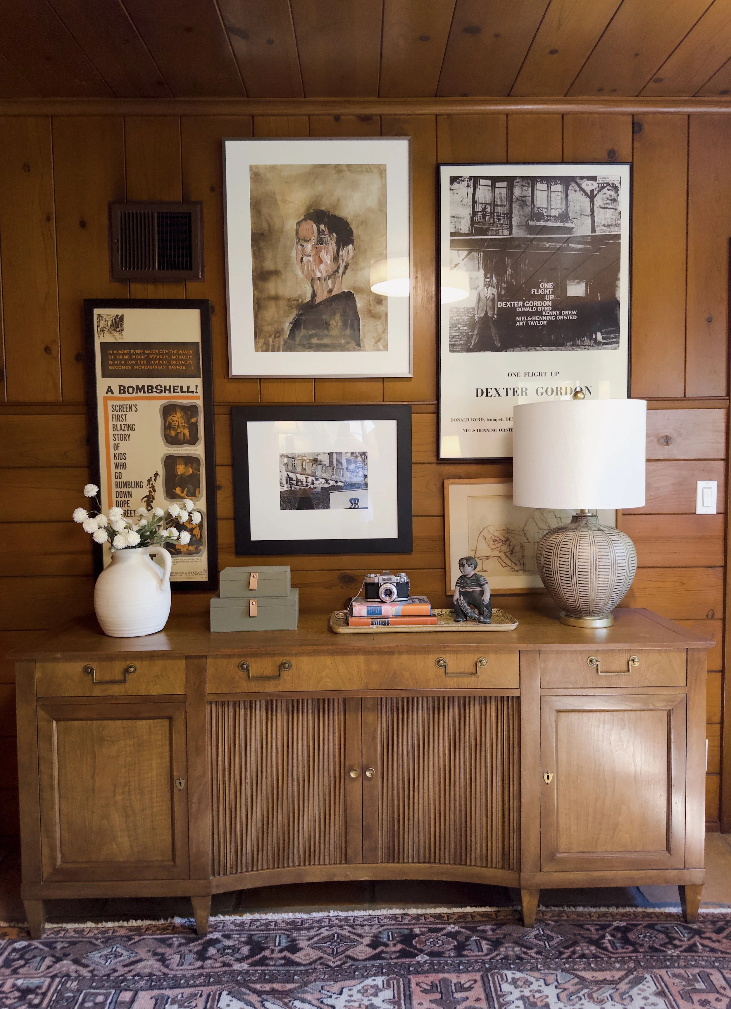 A wooden sideboard with framed pictures and artwork hanging on a wooden wall above it. A white pitcher with white flowers, a small stack of boxes, a vintage camera, and a sculpture are on the sideboard. A large white cylindrical lamp is on the right 
