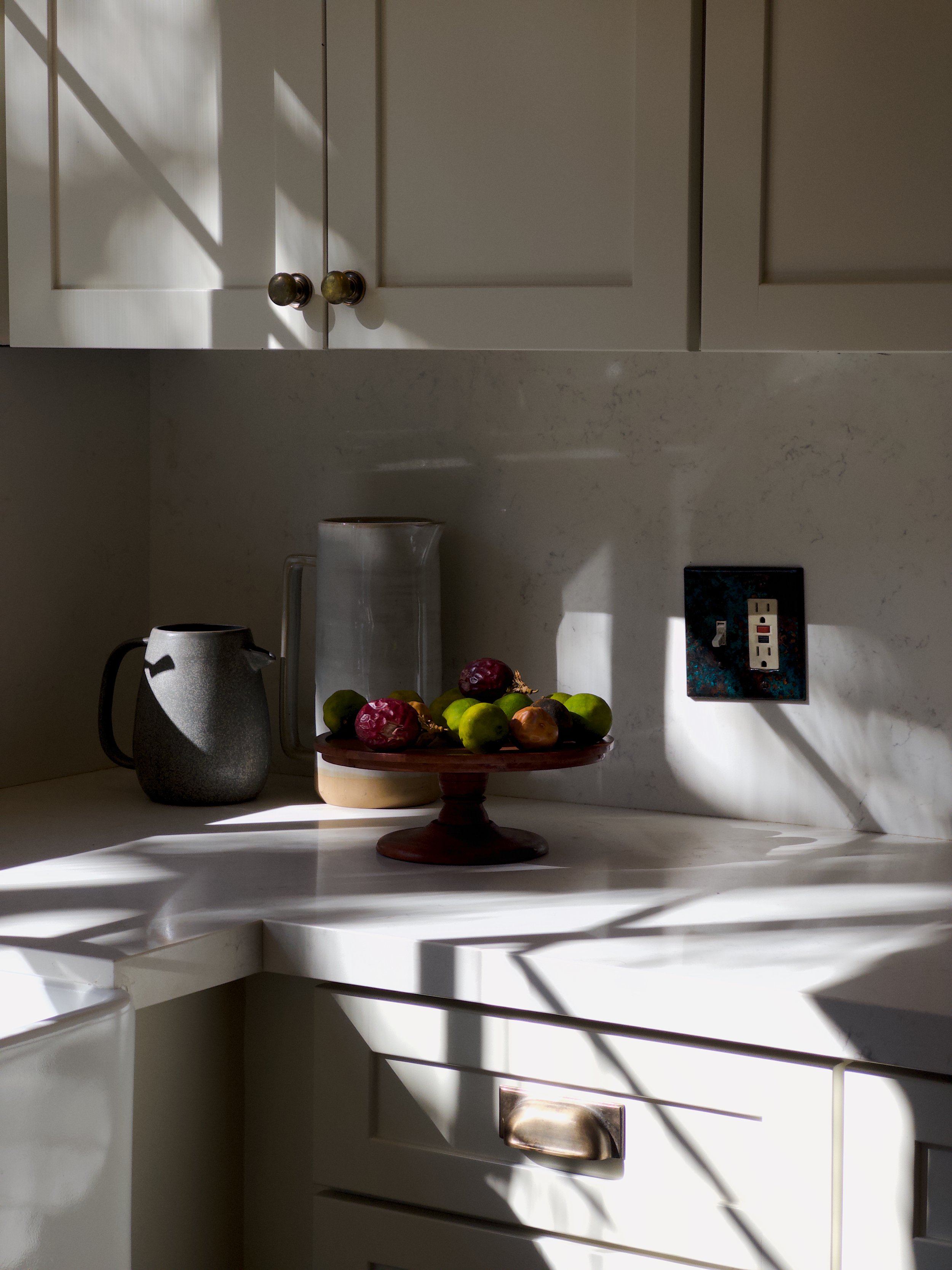 Kitchen countertop with a plate of apples, a ceramic coffee pot, a glass pitcher, and a power outlet, with sunlight casting shadows.