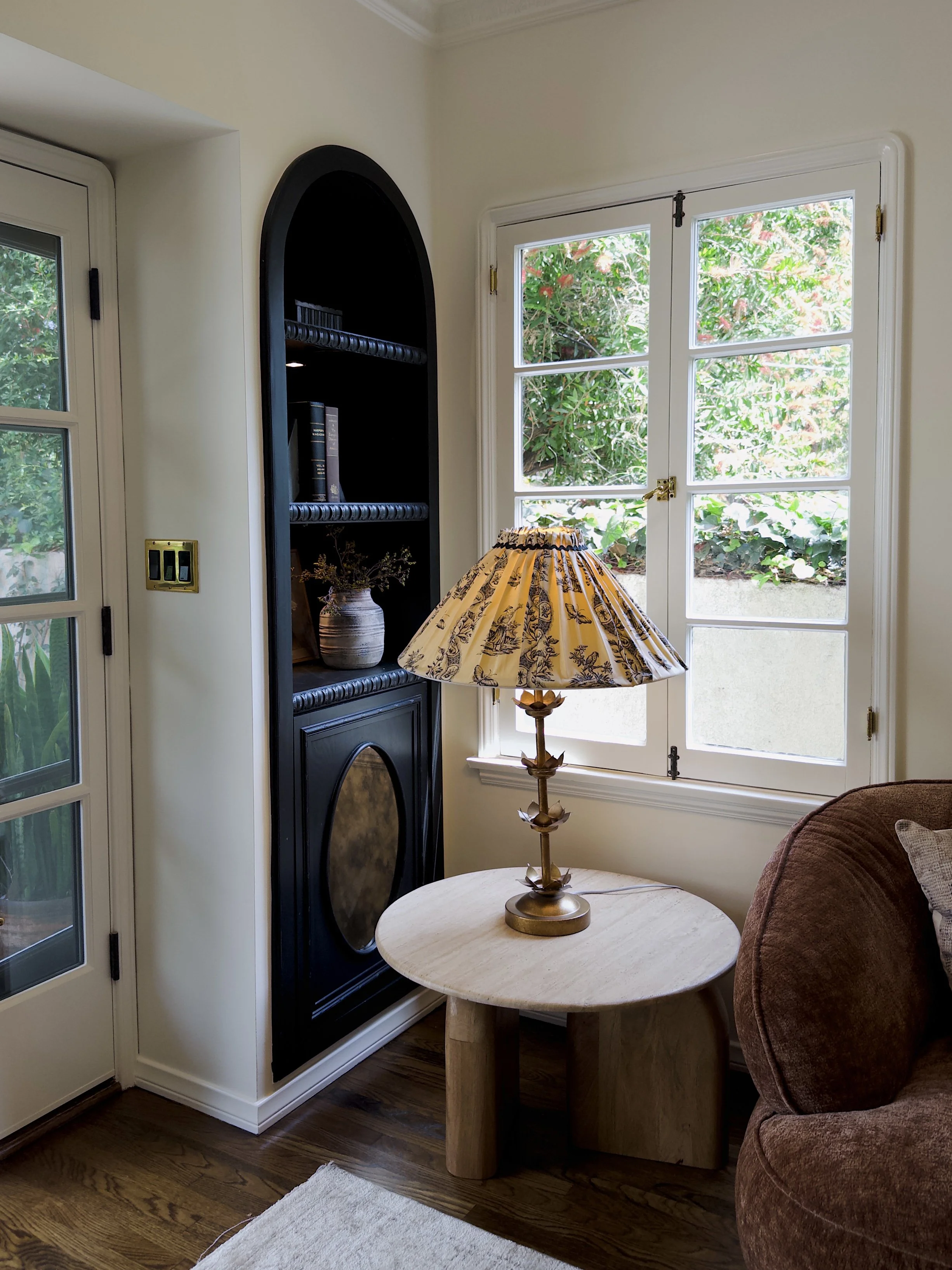A cozy living room corner featuring a beige round table with a decorative lamp, a brown upholstered armchair, a black built-in bookshelf with a vase and books, near double glass doors and a large window letting in natural light and showing green outd