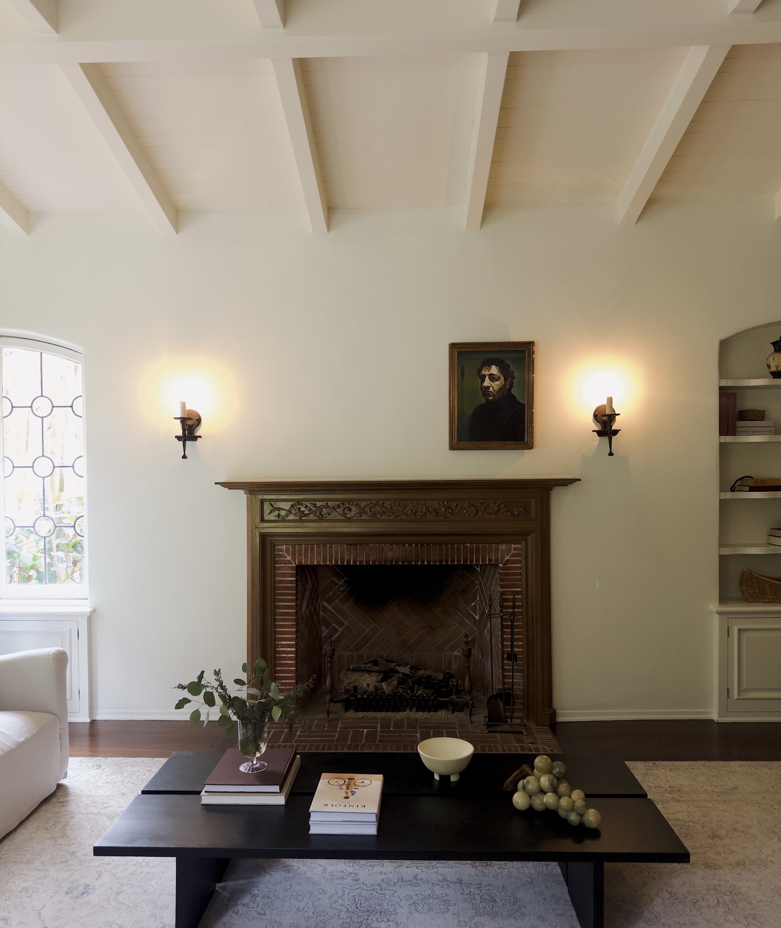 Living room with a brick fireplace, framed portrait above, two wall sconces, a black coffee table with books, a decorative bowl, and grapes, cream-colored walls, built-in shelves, and a window with decorative glass to the left.