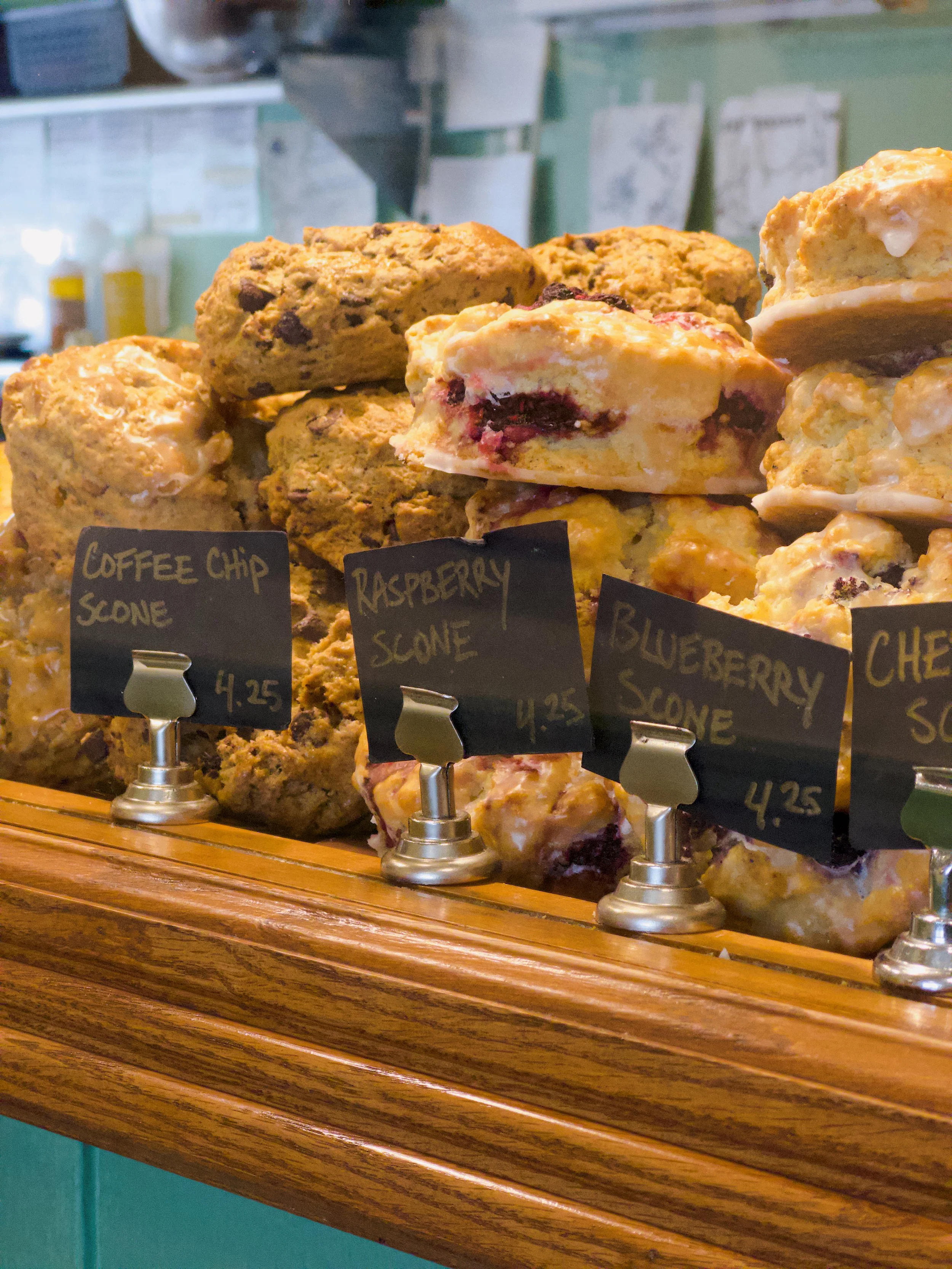 Display of various baked muffins, including coffee chip, raspberry, blueberry, and cherry flavors, with small black labels displaying their names and prices in a bakery or café setting.