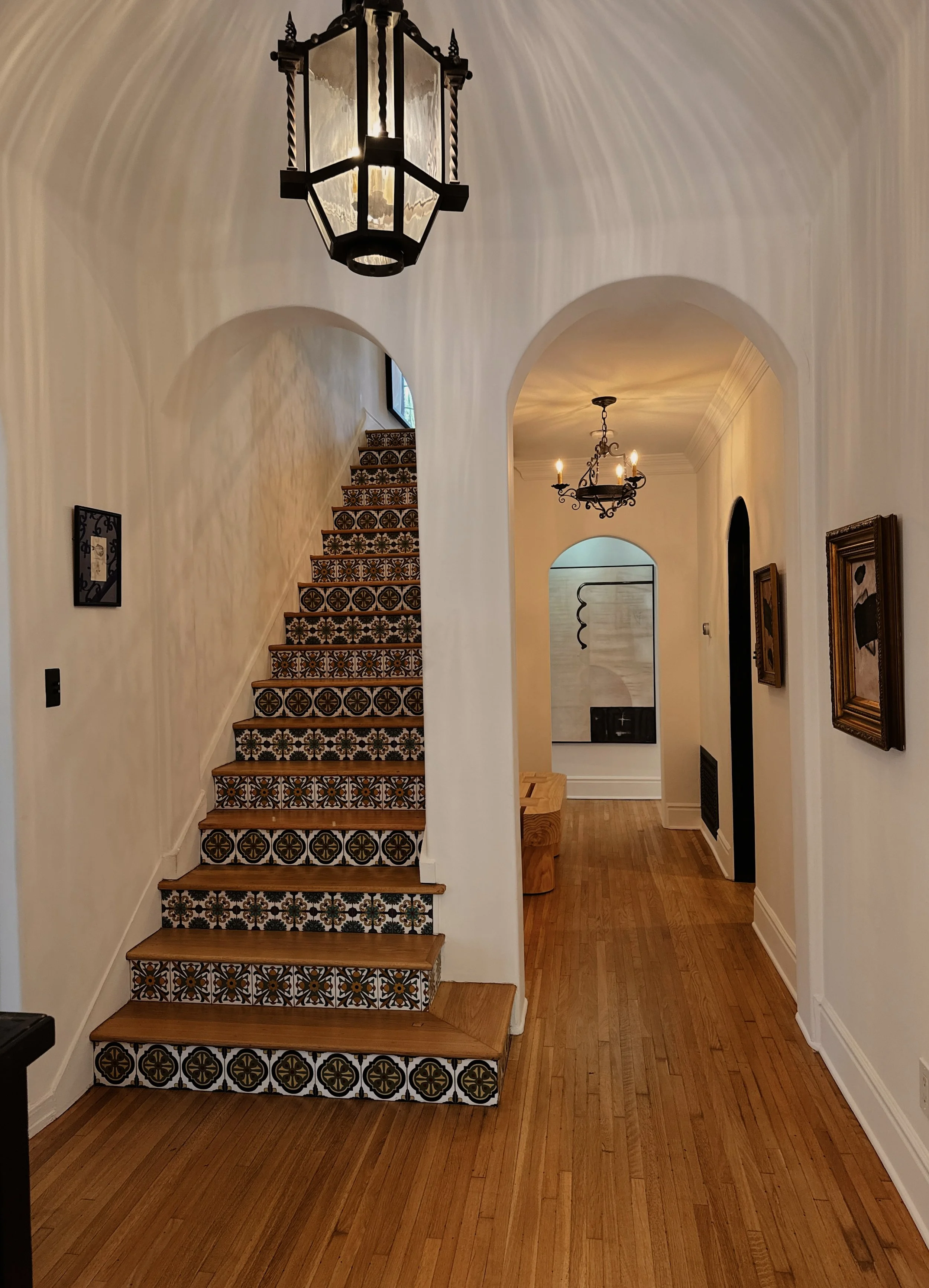Interior view of a hallway with a staircase featuring decorative patterned tiles on the risers, arched doorways, wooden flooring, and ornate chandeliers hanging from the ceiling.