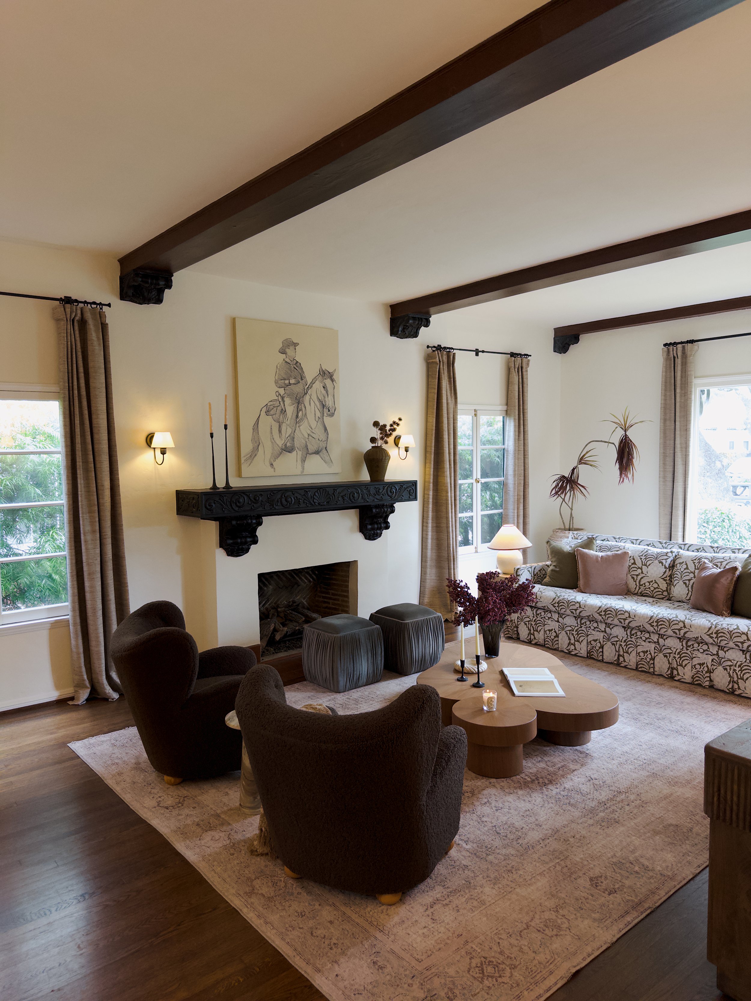 A cozy living room with a sofa, armchairs, a coffee table, and a fireplace with a mantel. There are large windows with curtains, a painting of a person on a horse above the fireplace, and decorative plants. The room features dark wood beams on the ce