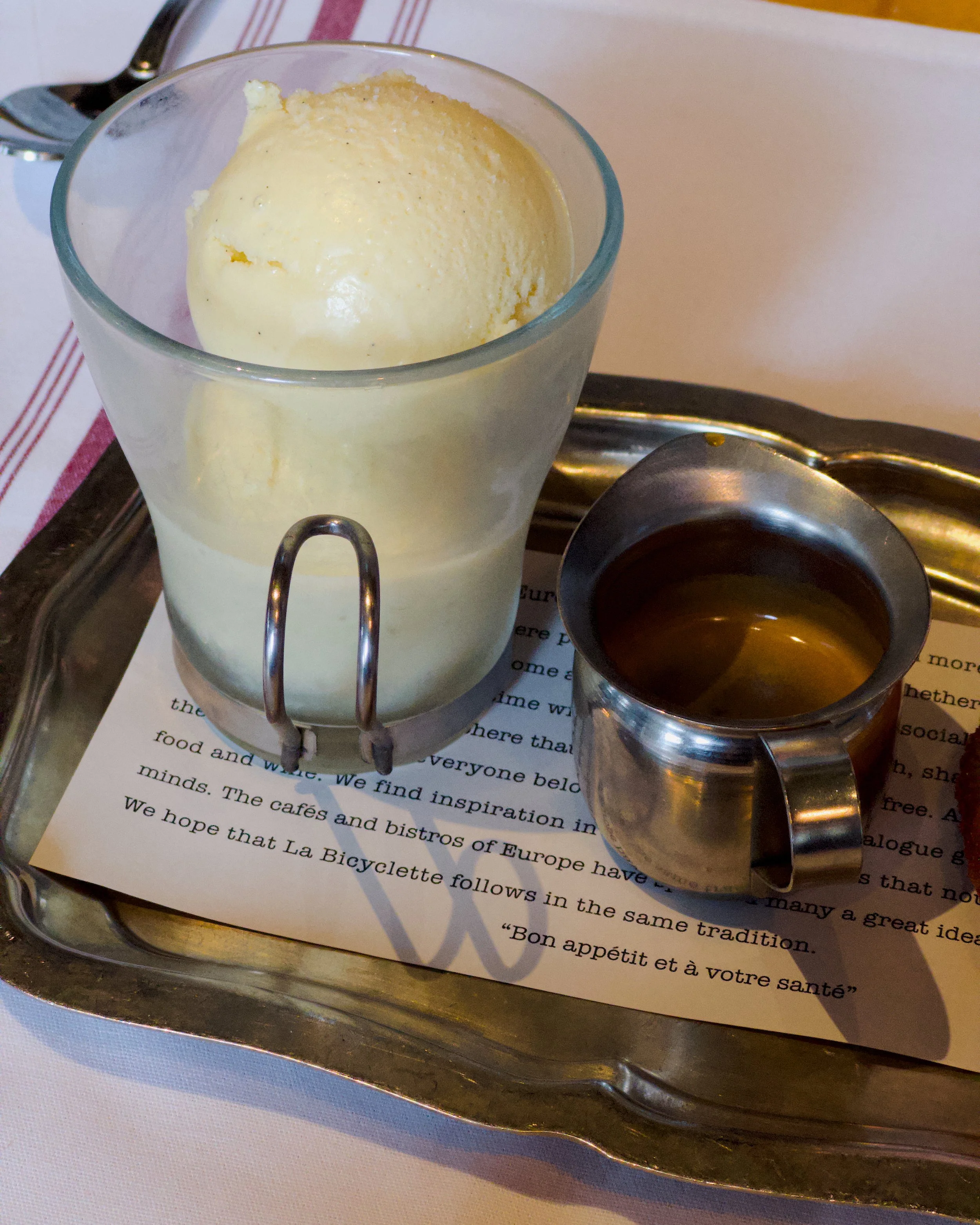 A glass of vanilla ice cream on a metal tray with a small pitcher of chocolate syrup and a fork.