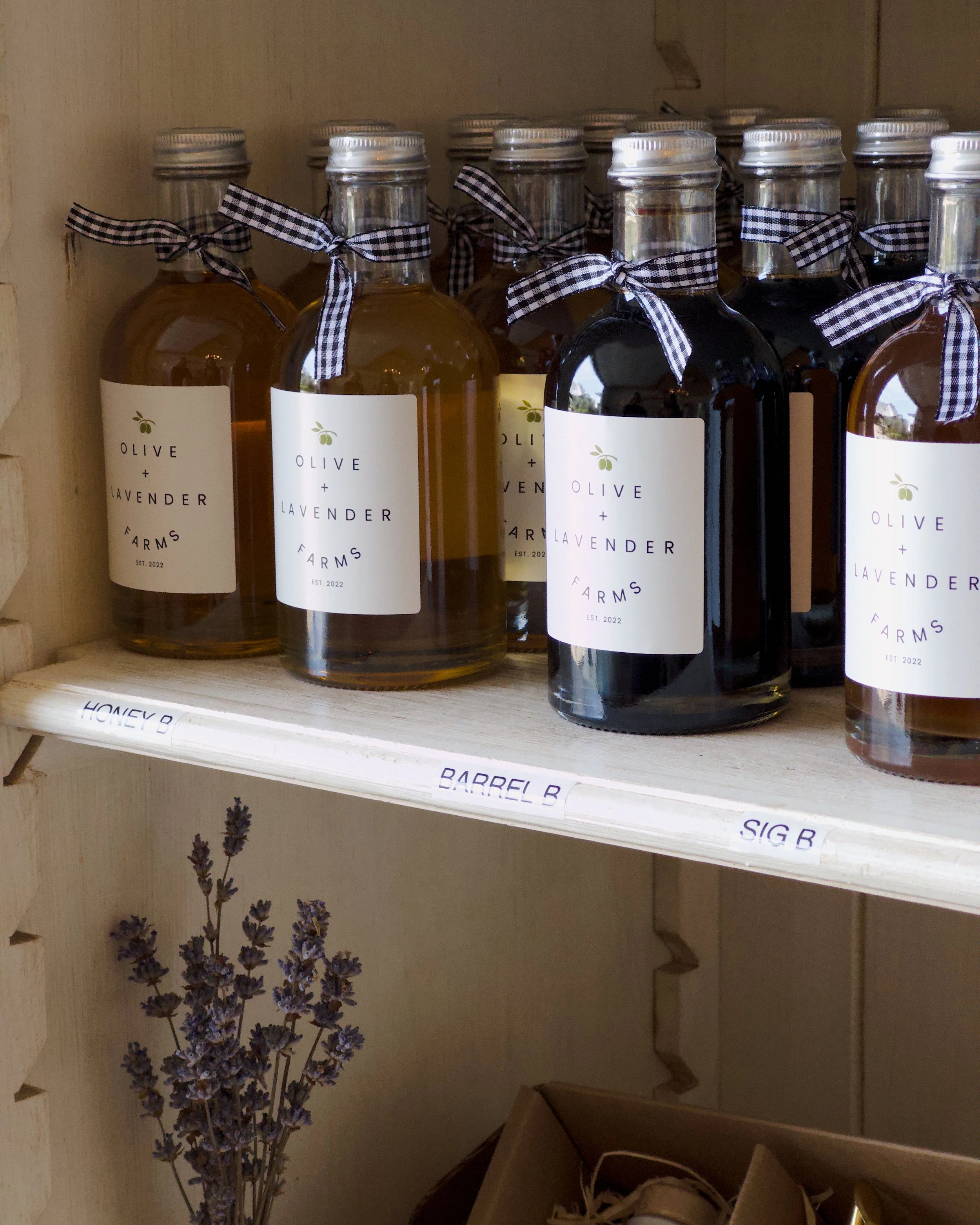 Several glass bottles filled with liquid, tied with black and white checkered ribbon around the neck, labeled 'Olive + Lavender Farms EST. 2022,' on a wooden shelf with lavender flowers below.