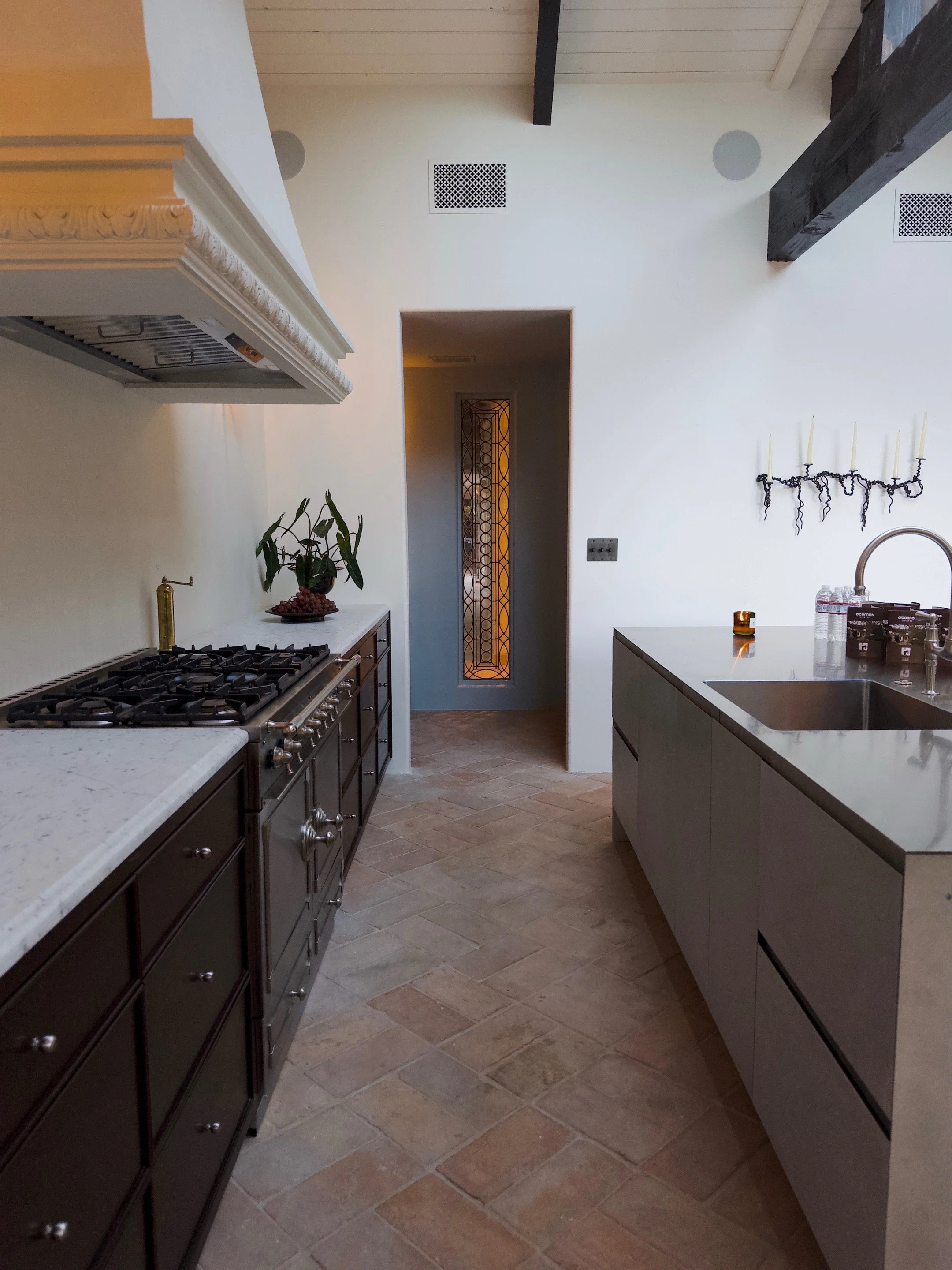 A kitchen with a white countertop, dark lower cabinets, a stove, a potted plant, a framed glass door, and a wall-mounted candle holder with six unlit candles.