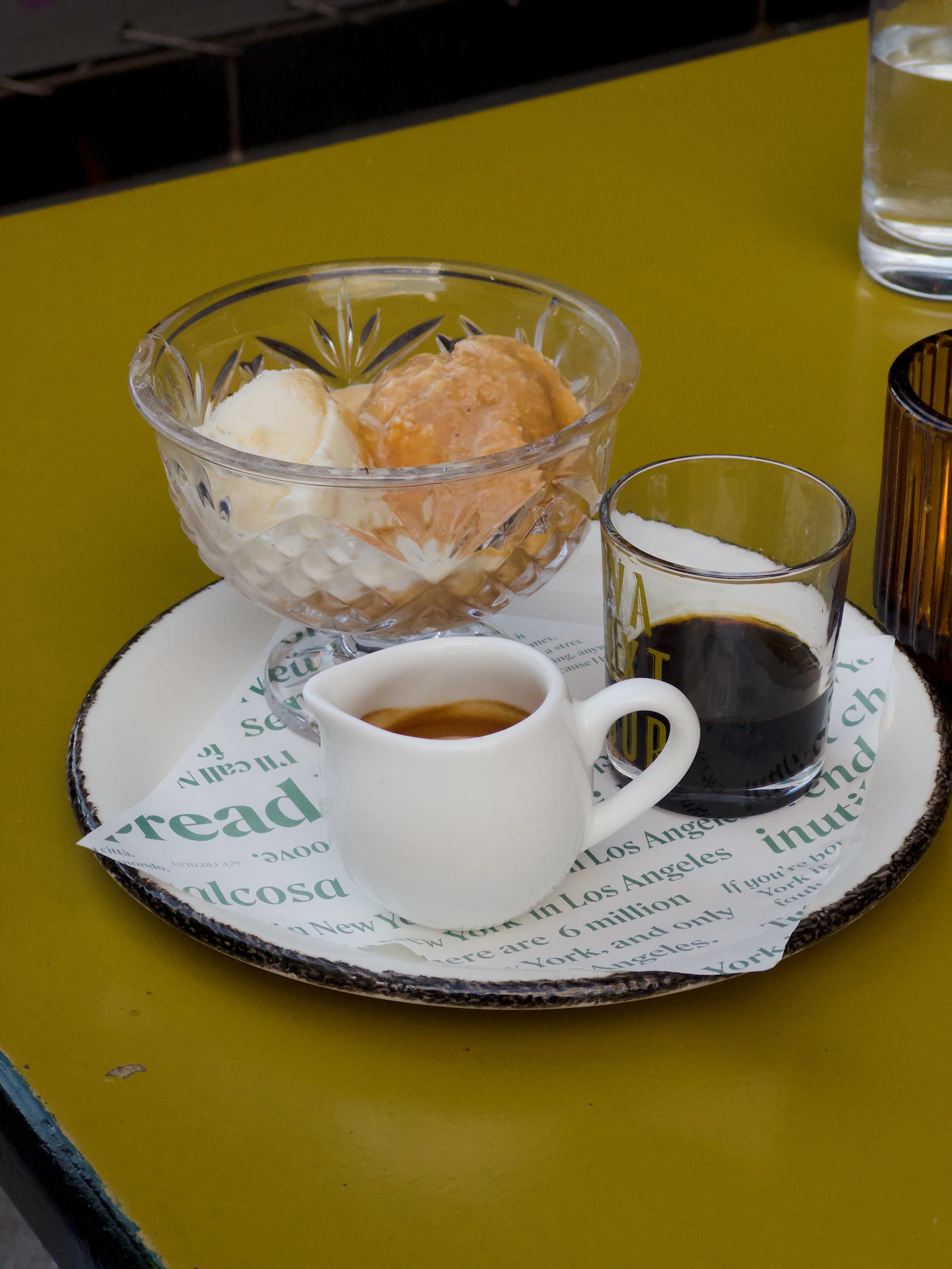 A glass bowl containing two scoops of ice cream, one vanilla and one caramel. The bowl is on a tray with a paper liner, along with a cup of espresso, a glass of water, and a small jug of cream.