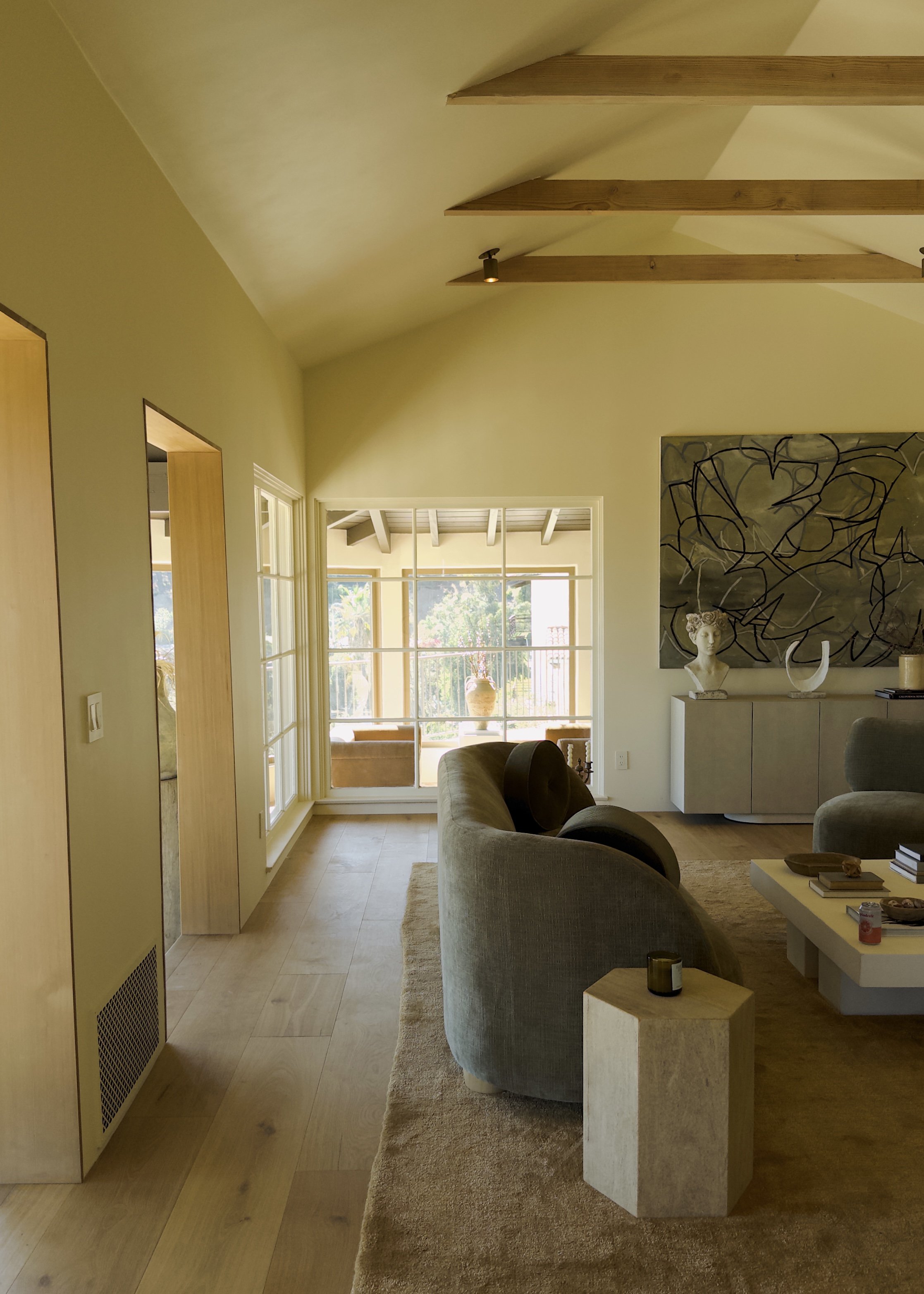 Living room with beige walls, large window, wooden beams on ceiling, a modern abstract wall art, a bust sculpture, beige sofa, coffee table with books, and small decor items.