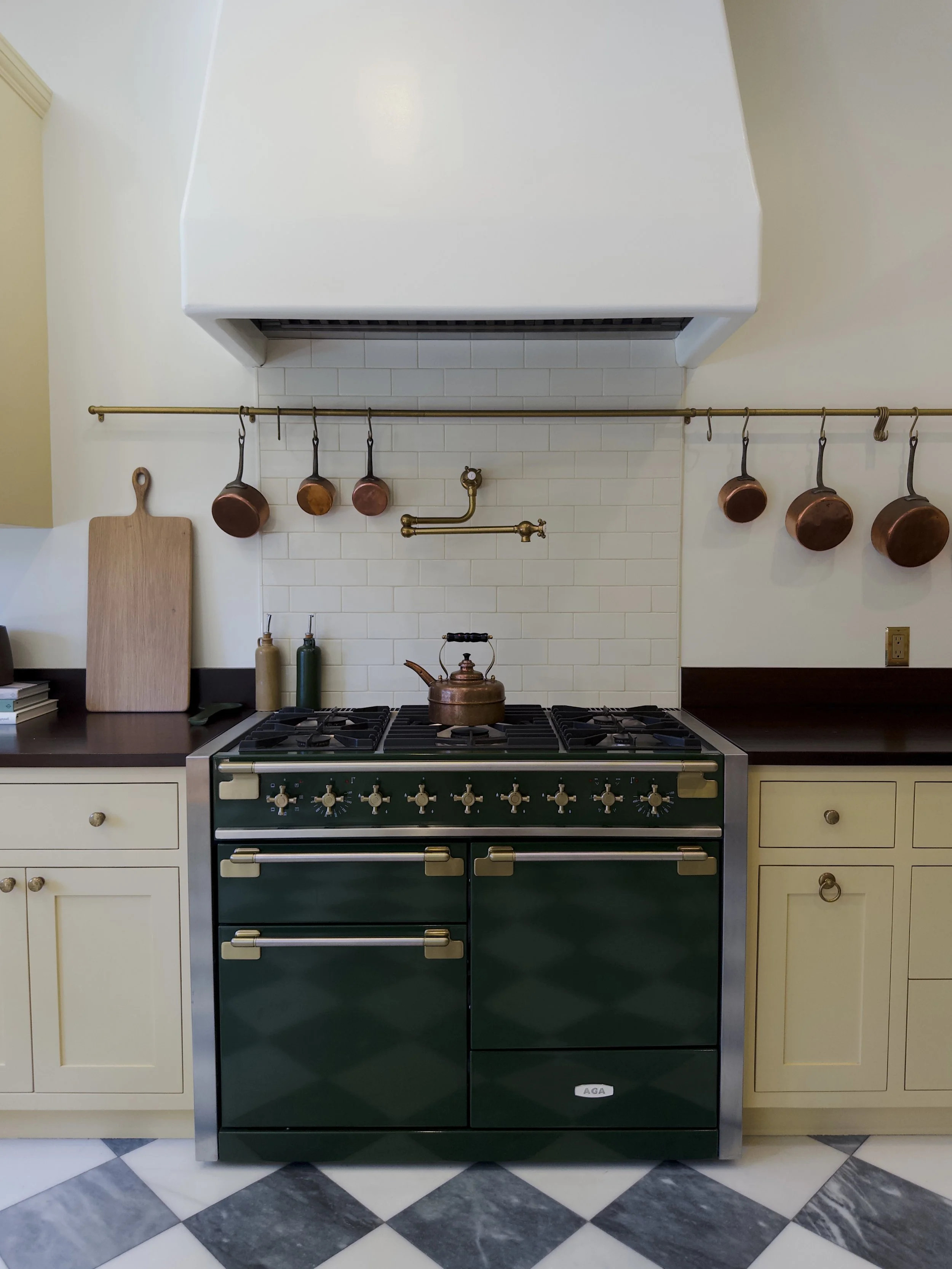 A kitchen with a dark green oven, white cabinets, hanging copper pots, a wooden cutting board, and a black and white checkered tile floor.