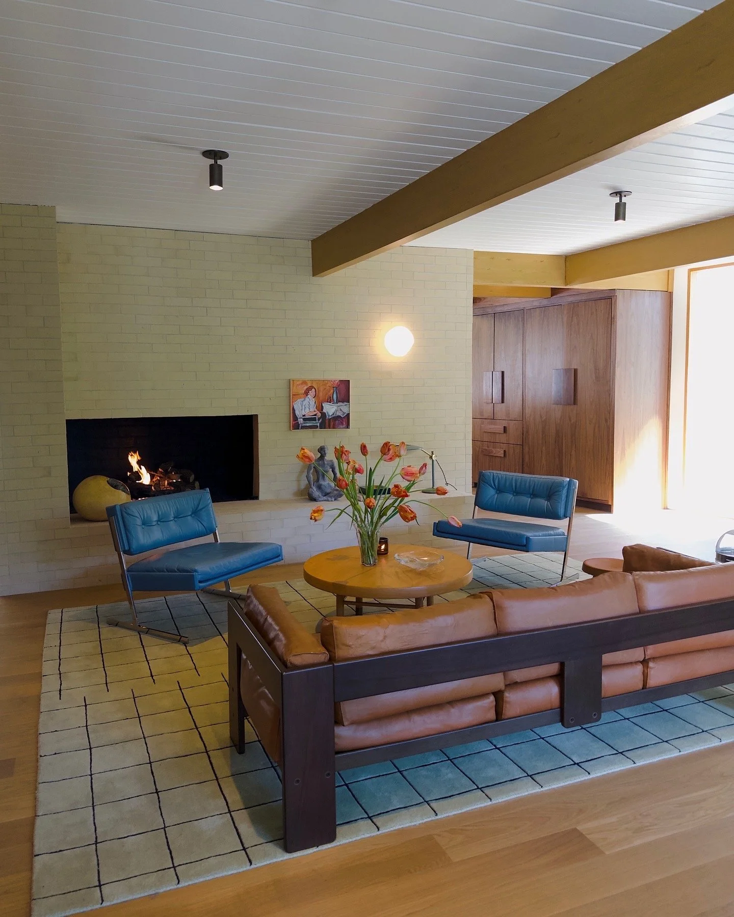 Living room with yellow brick wall, fireplace, blue chairs, brown leather couch, wooden coffee table with a vase of orange flowers, wall art, and a wooden cabinet. The room has a white ceiling with wooden beams and natural light coming from a large window.