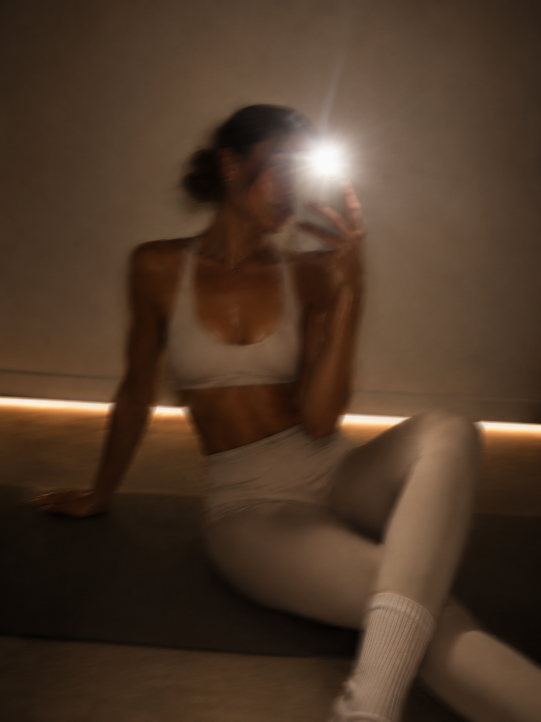 A woman taking a mirror selfie with her phone, wearing a white sports bra and leggings, sitting on the floor in a room with minimalistic decor and soft lighting.