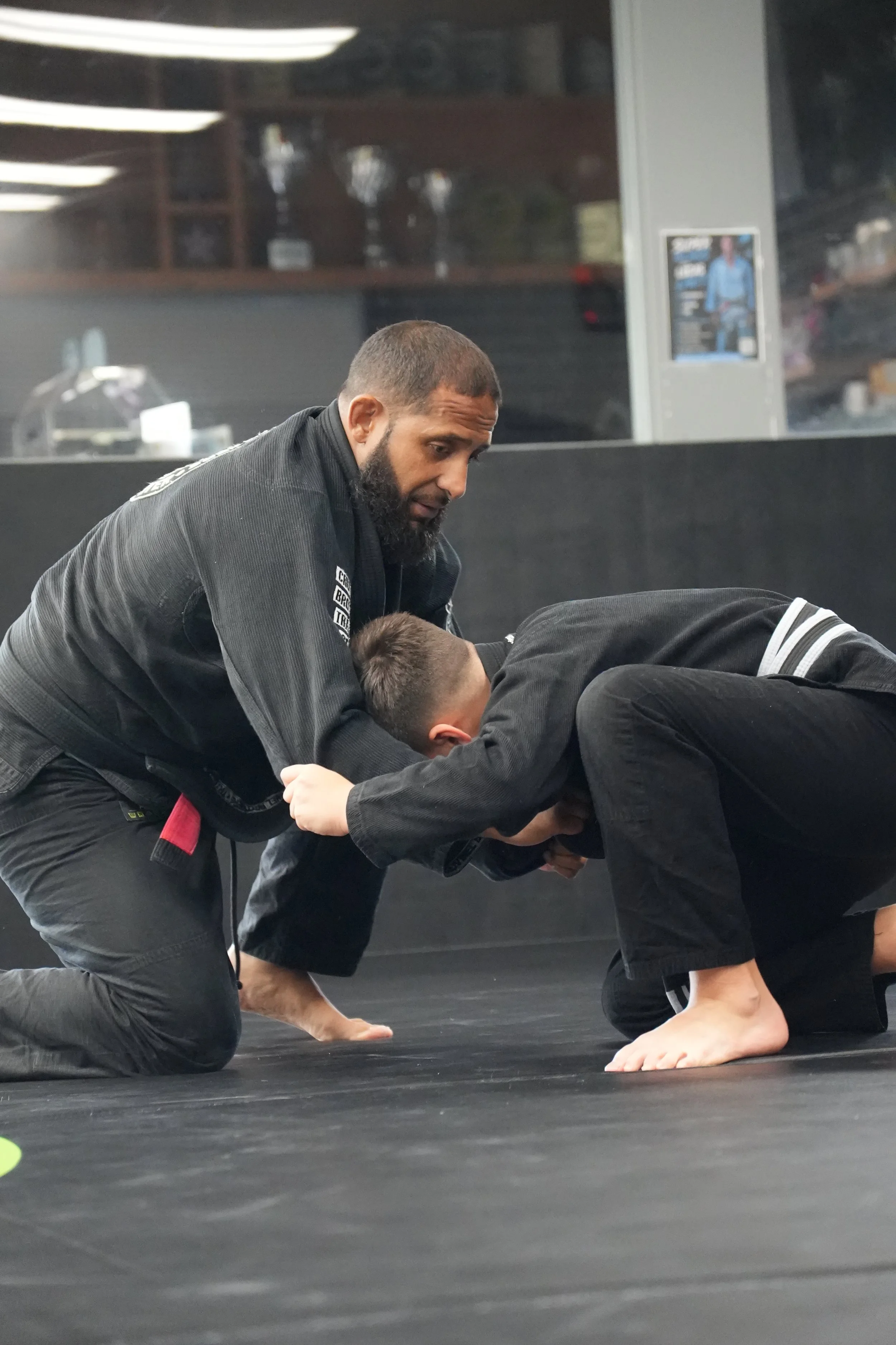 A man with a beard and a black martial arts gi is helping a young person in a black gi practice a grappling move on a black mat in a martial arts training facility.