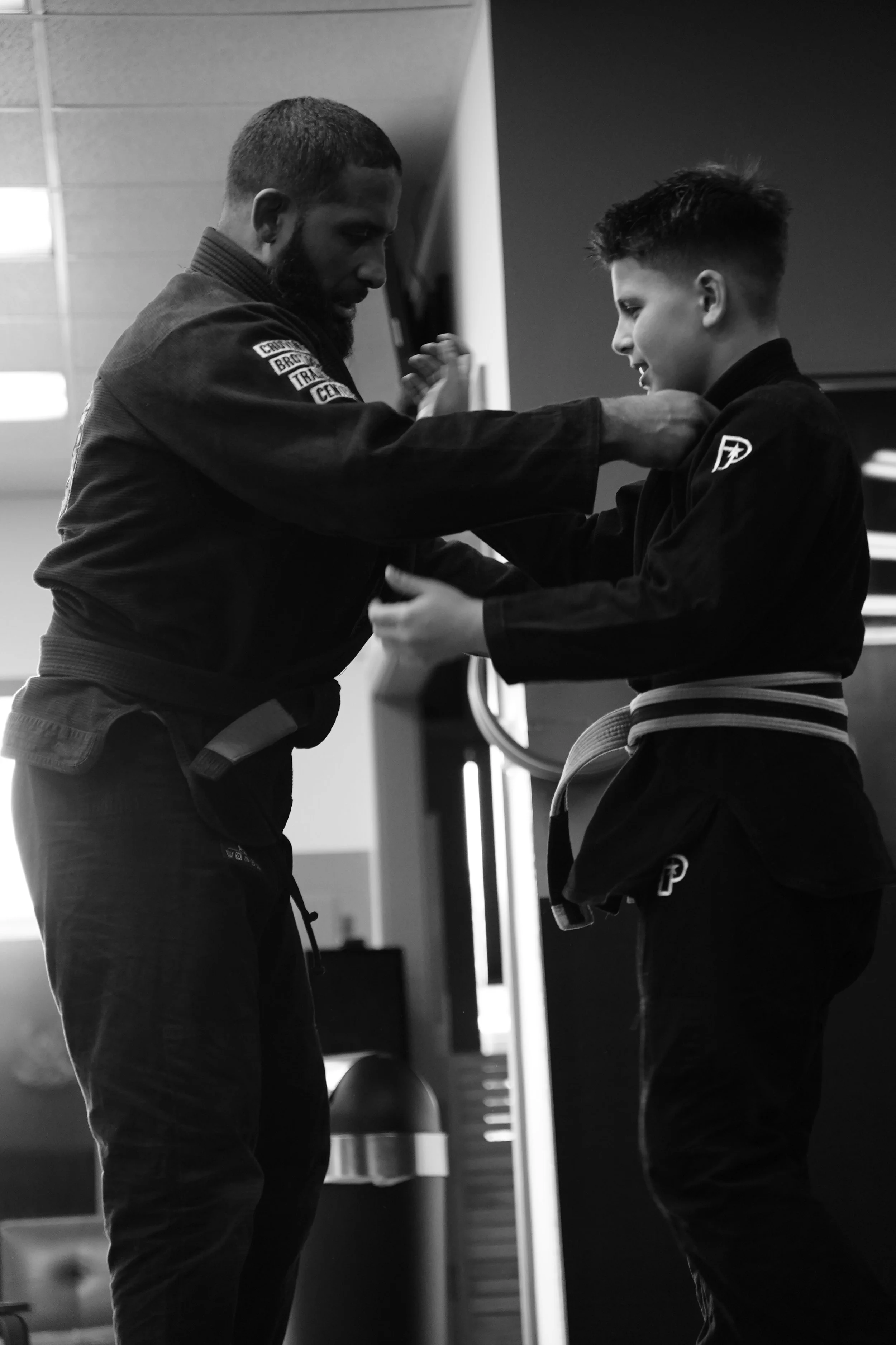 A black and white photo of an instructor training a young student in martial arts, both in gi uniforms, inside a gym or dojo.