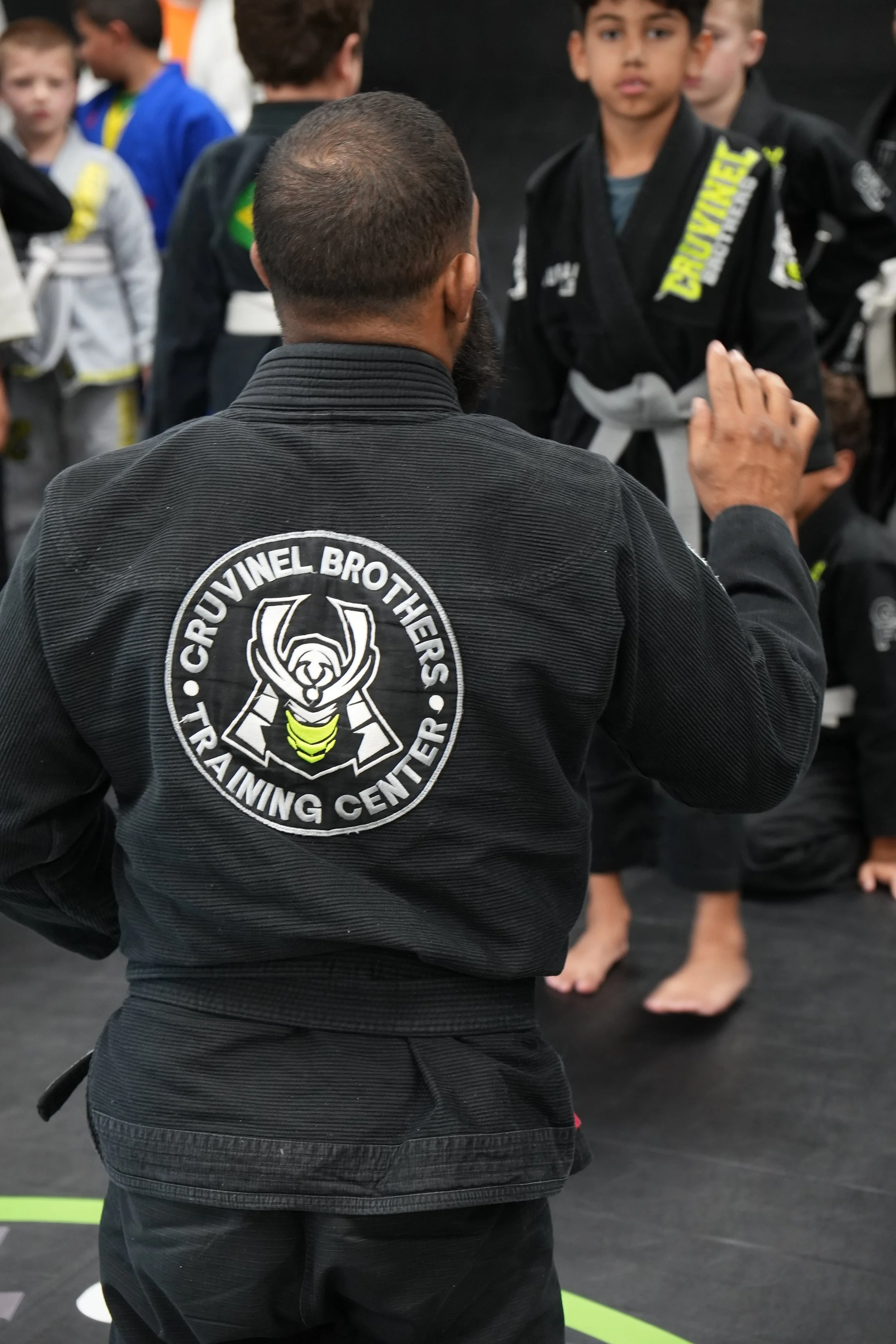 A man wearing a black Brazilian Jiu-Jitsu gi with a patch from the Groovinel Brothers Training Center on the back, standing with his back to the camera in a dojo. Several children, also in gi uniforms, are standing in the background.