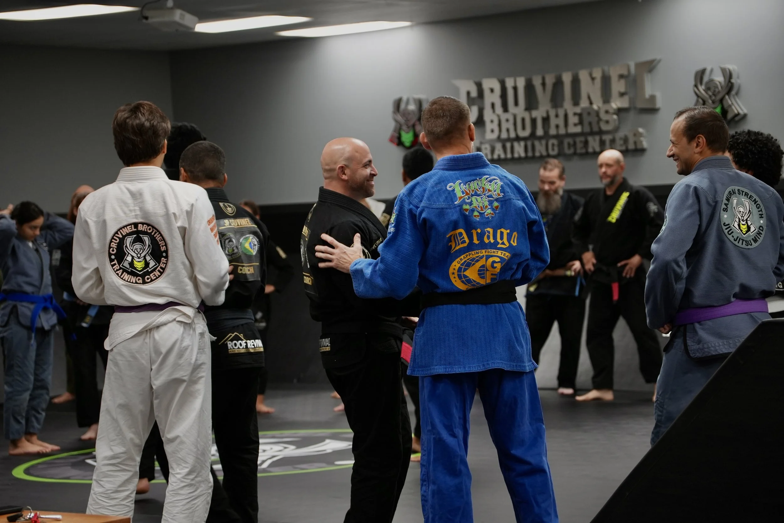 Brazilian Jiu-Jitsu practitioners wearing gis with various patches, some smiling and shaking hands, inside a training center with a large logo on the wall reading 'Cruyinel Brothers Training Center.'