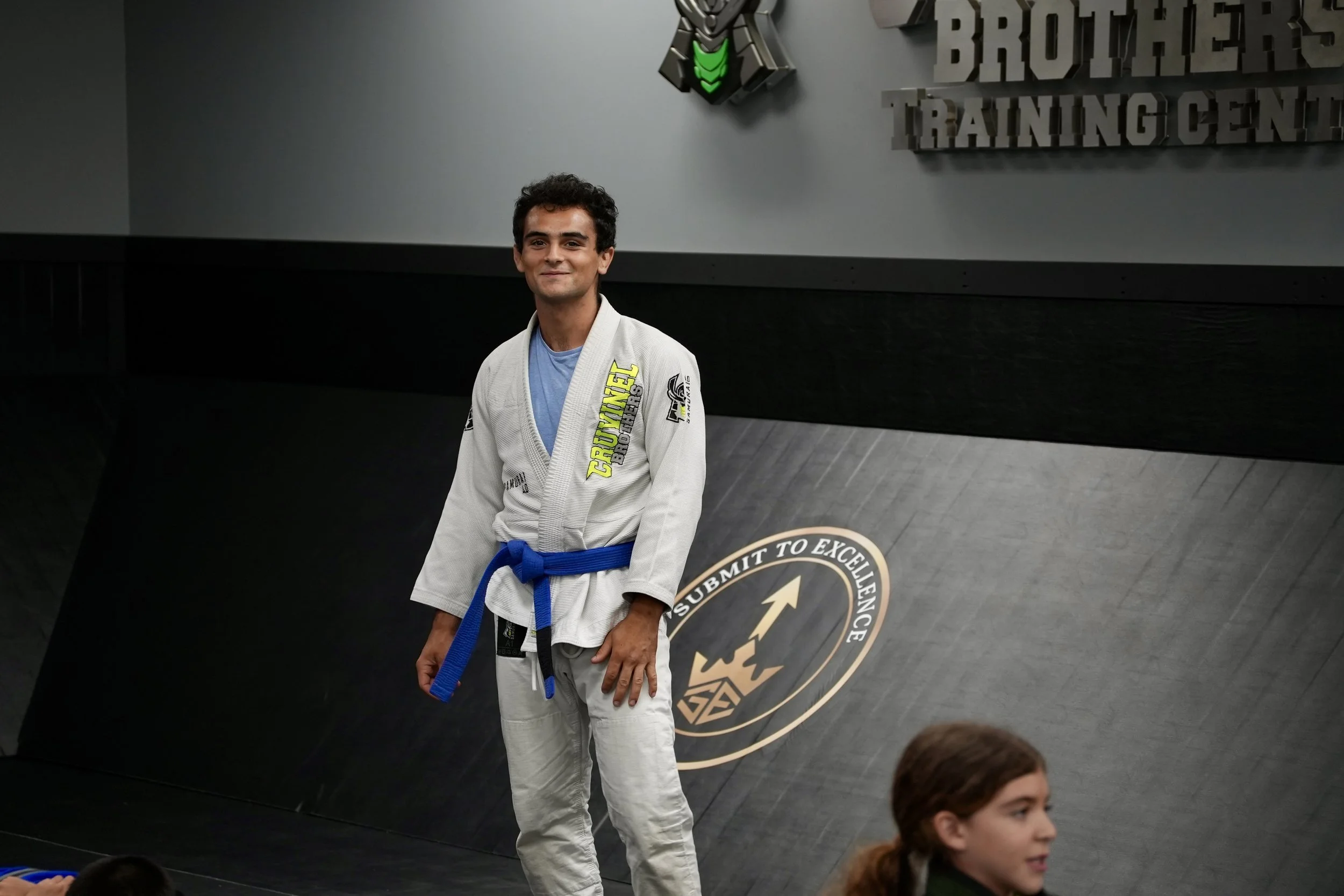 A man in a white Brazilian Jiu-Jitsu gi with a blue belt standing in a martial arts training center. The background includes a wall with a logo and text about training, along with other children sitting on the floor.