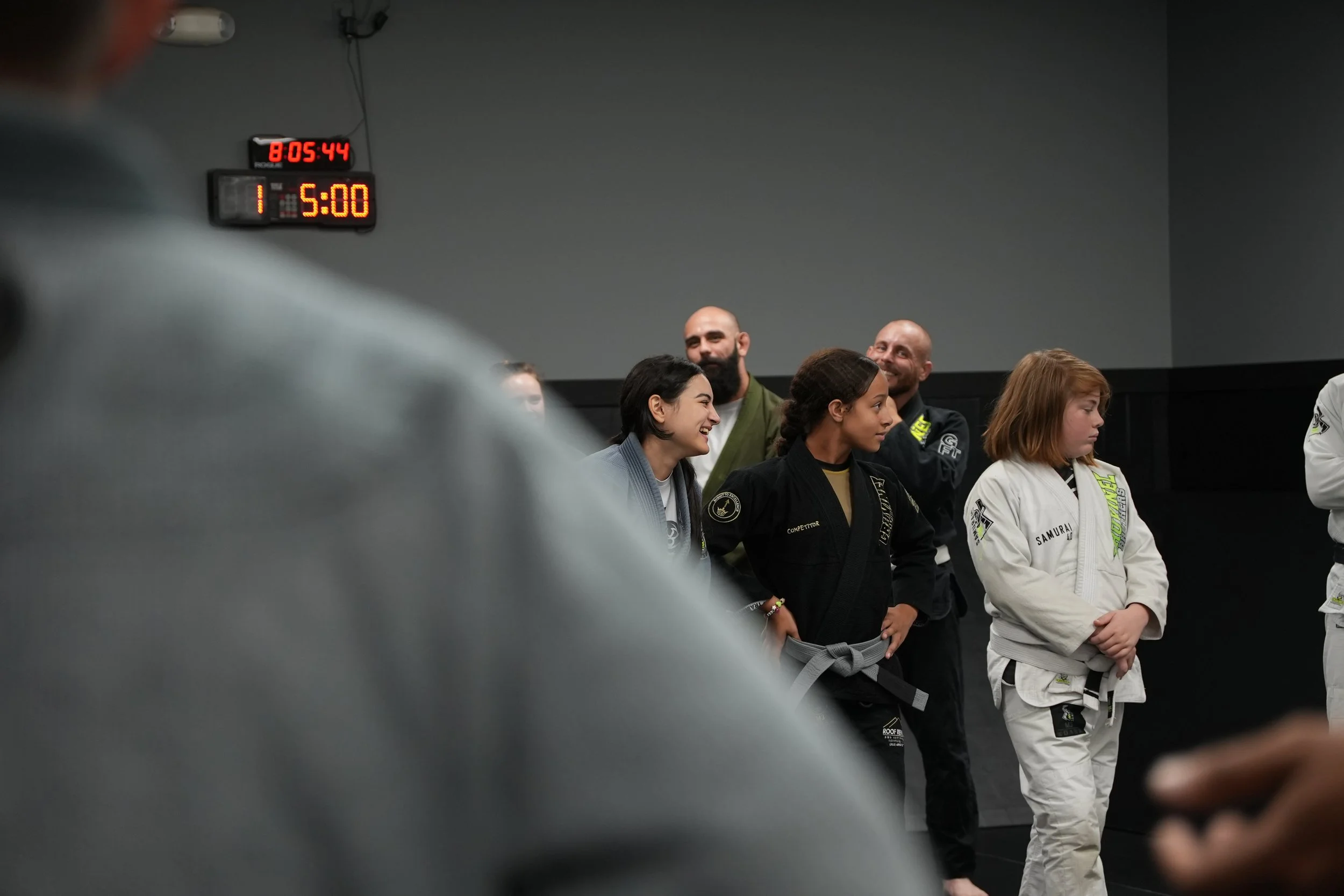 A group of people, including women and men in martial arts uniforms, standing in a line in a martial arts studio, smiling and talking. A digital clock on the wall shows 8:05:44, and another timer indicates 1 hour and 5 minutes.