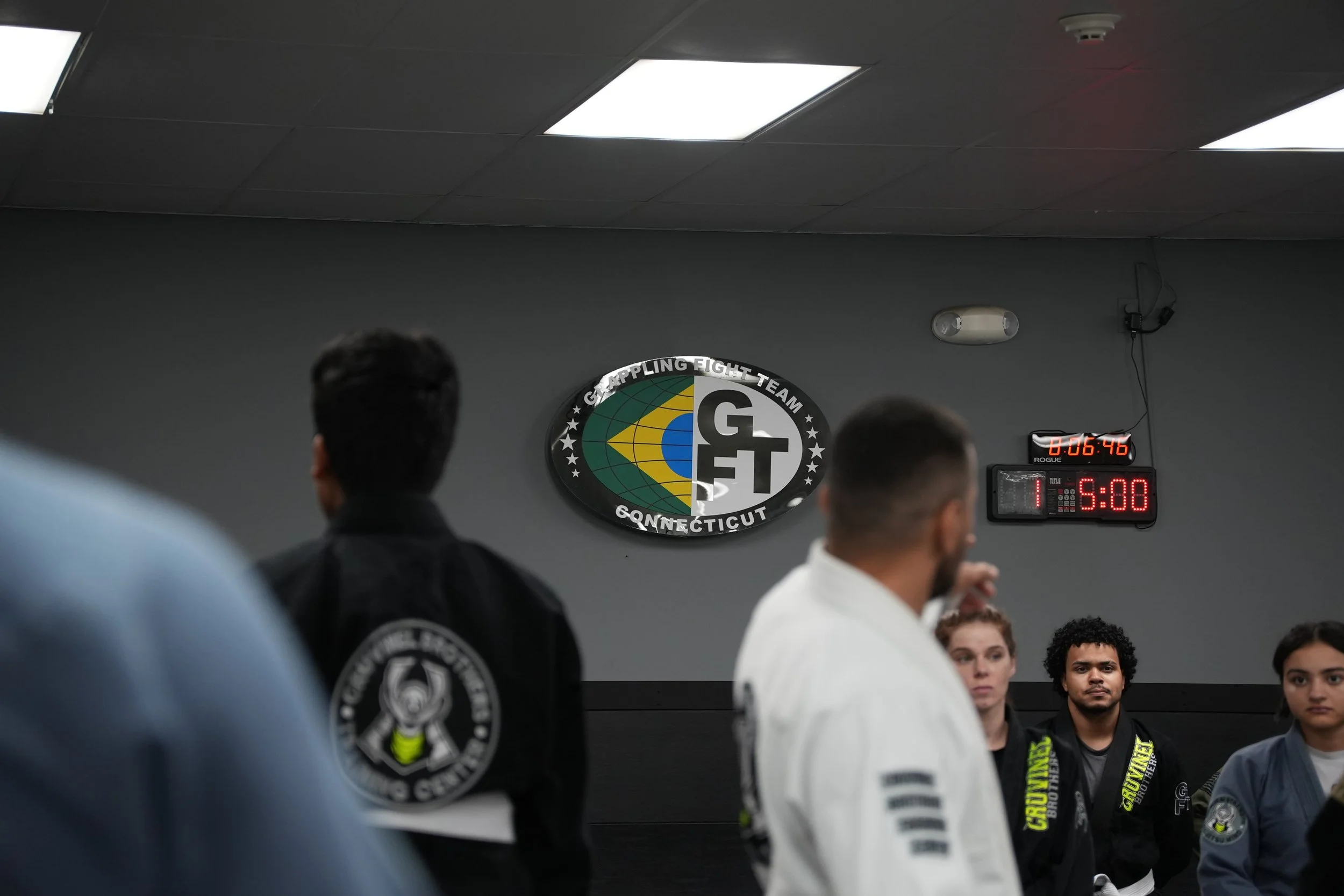 People participating in a martial arts class at Crosspointe Fight Team in Connecticut, with a logo on the wall and a timer, among students in martial arts uniforms.